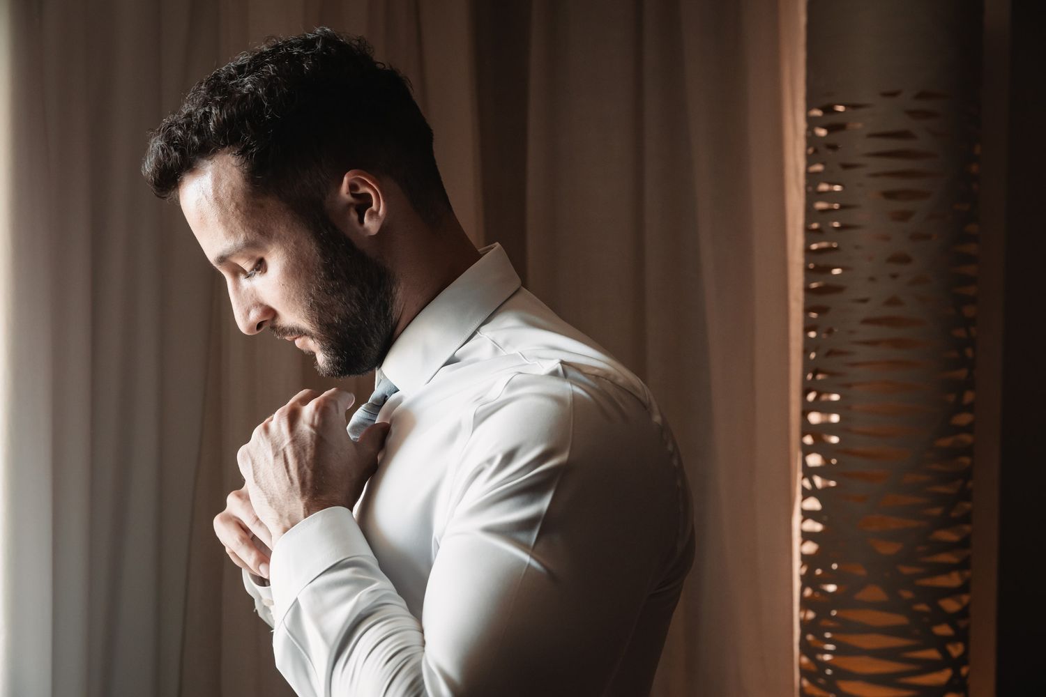 Elegant groom in white dress shirt adjusting his tie in a softly lit room before the wedding ceremony.