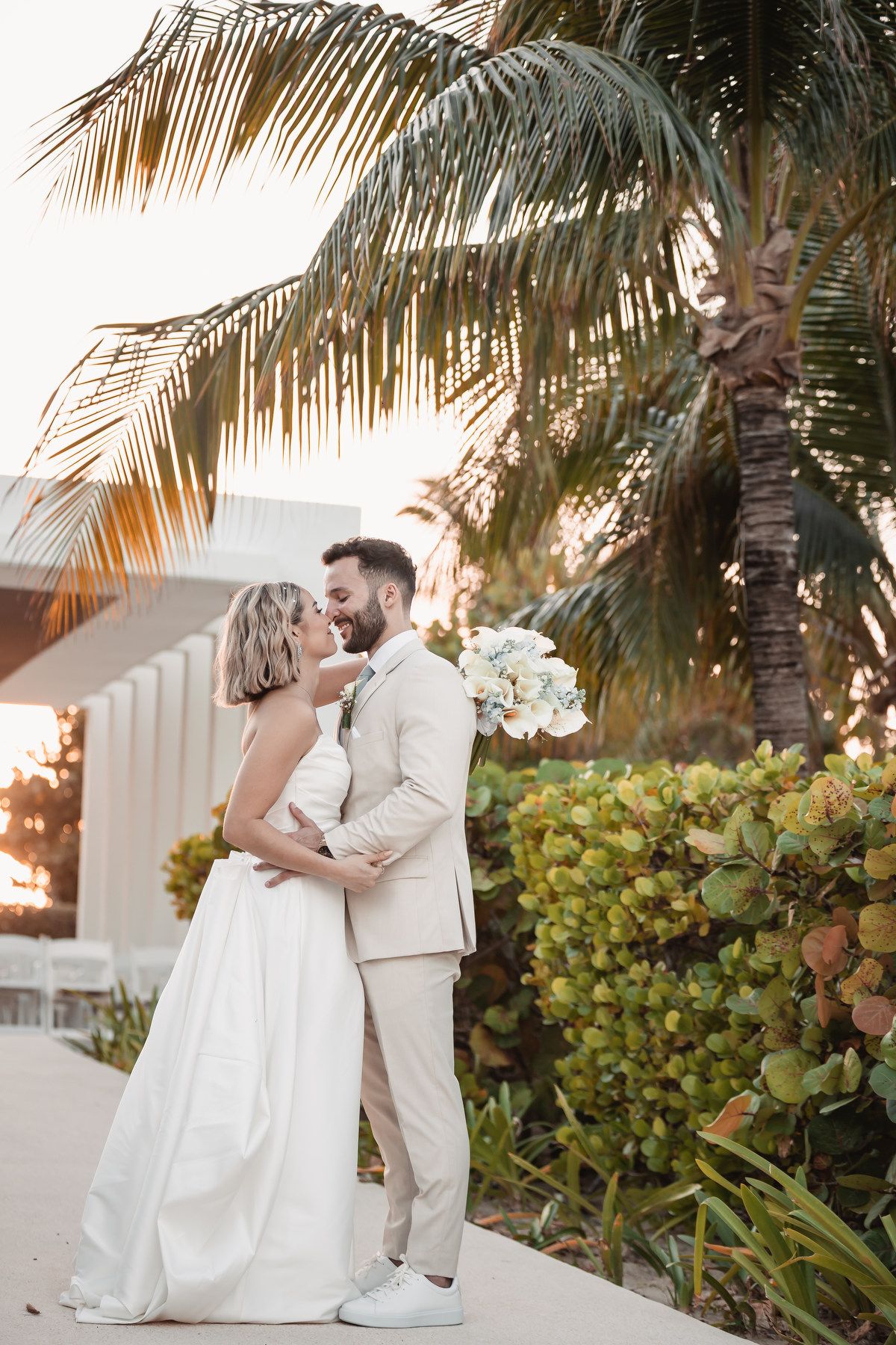 Bride and groom share romantic kiss under palm trees at golden hour sunset.