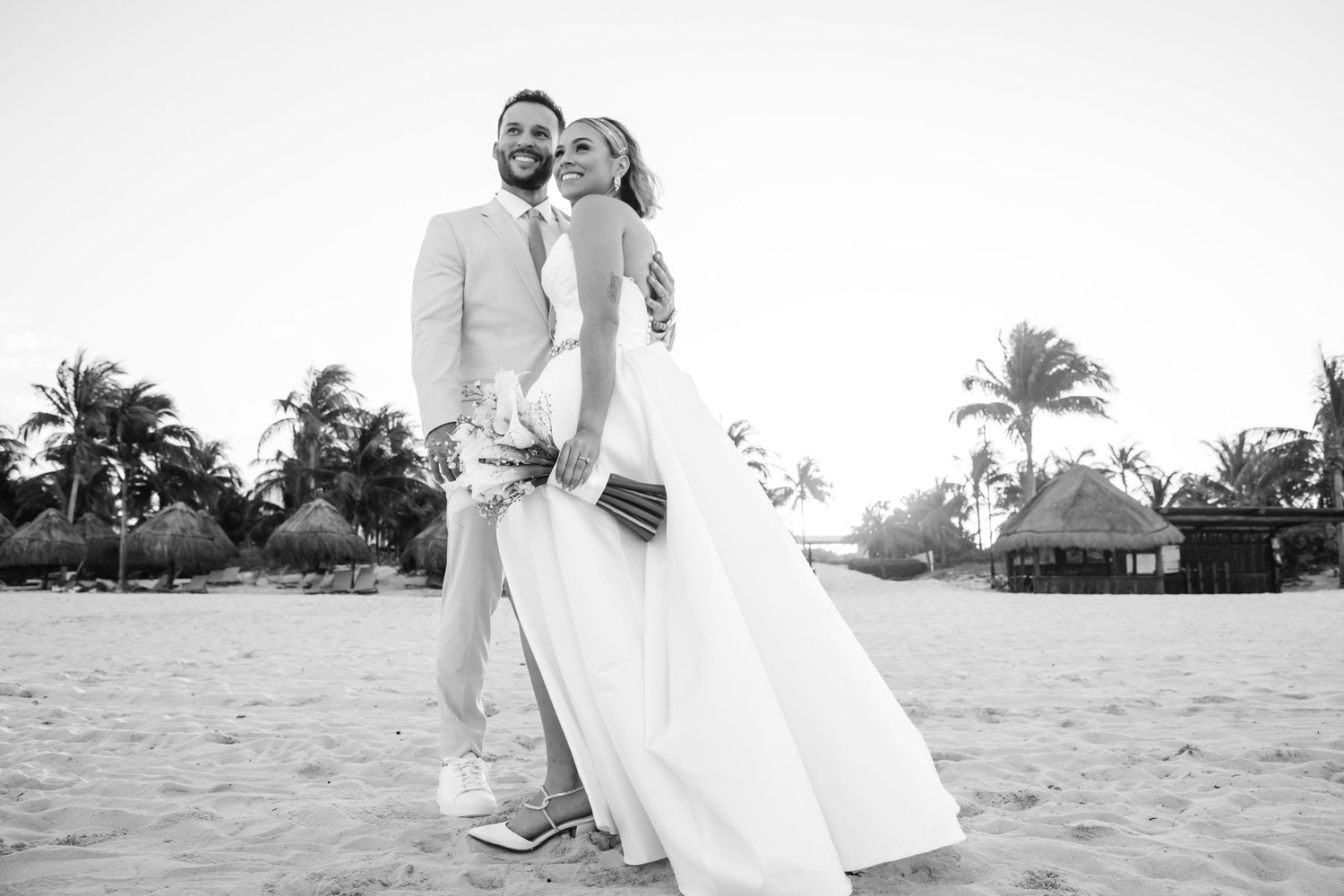 Black and white photo of newlyweds embracing on tropical beach with palm trees at destination wedding.