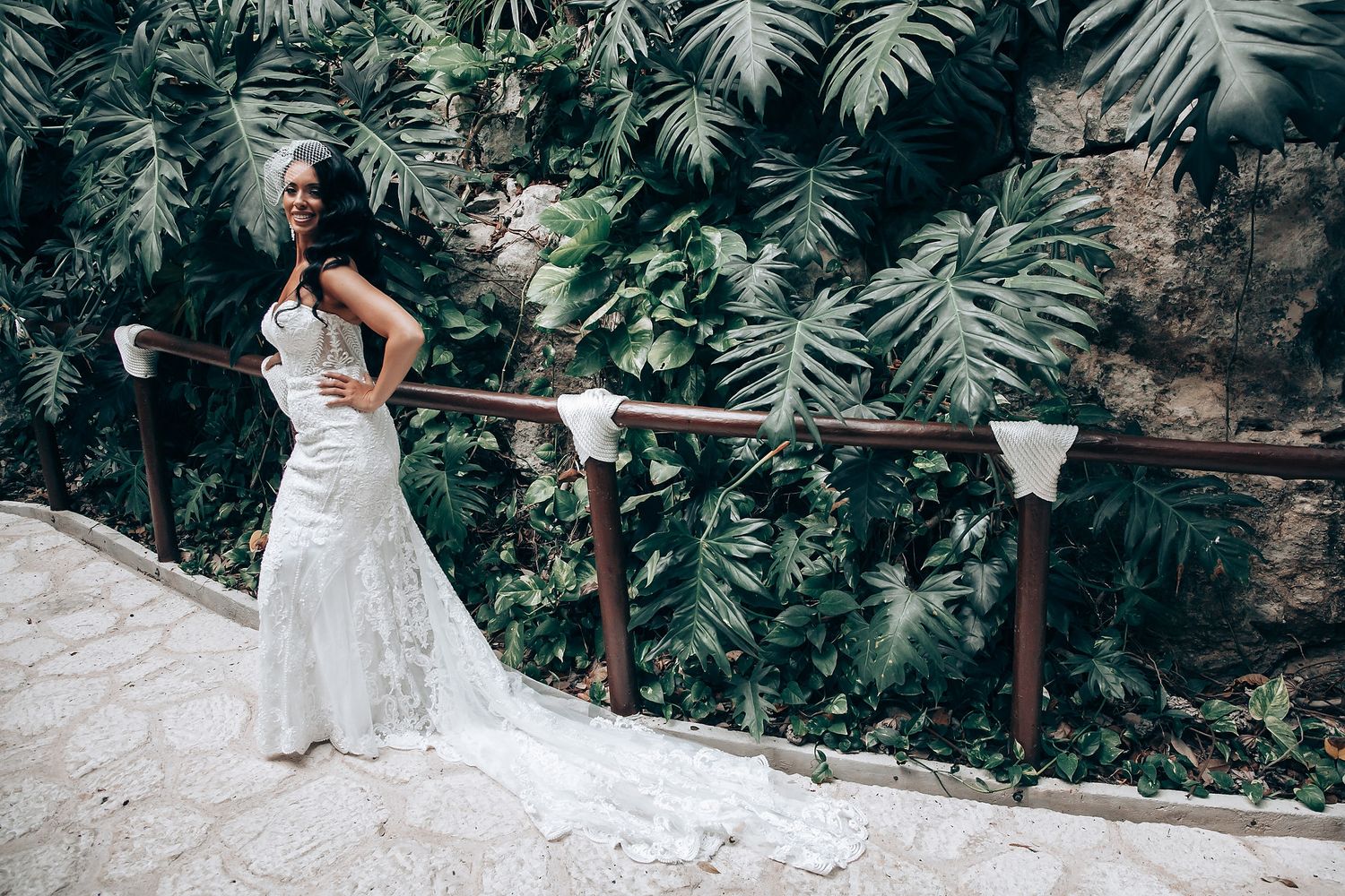 A bride poses on a stone pathway with tropical plants and wooden railing in the background.