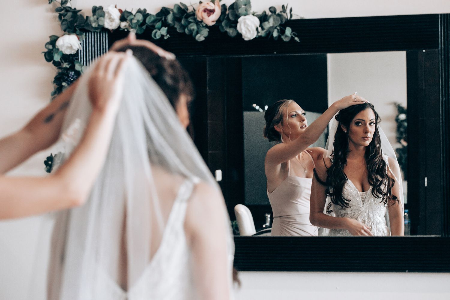 A bride in a white wedding dress adjusts her veil while looking in a mirror during wedding preparations.