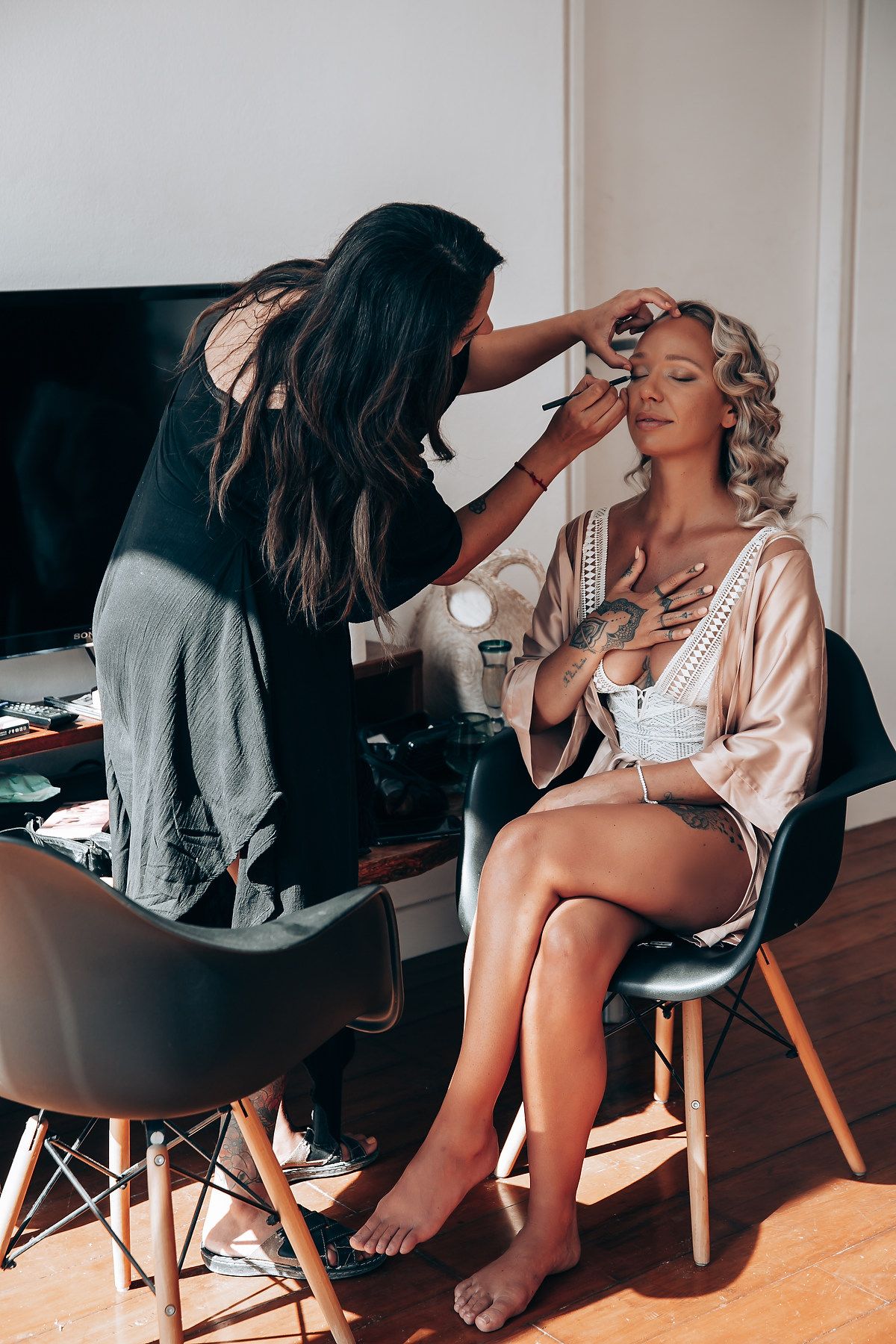 A makeup artist applies cosmetics to a model wearing a silk robe while sitting in a modern chair.