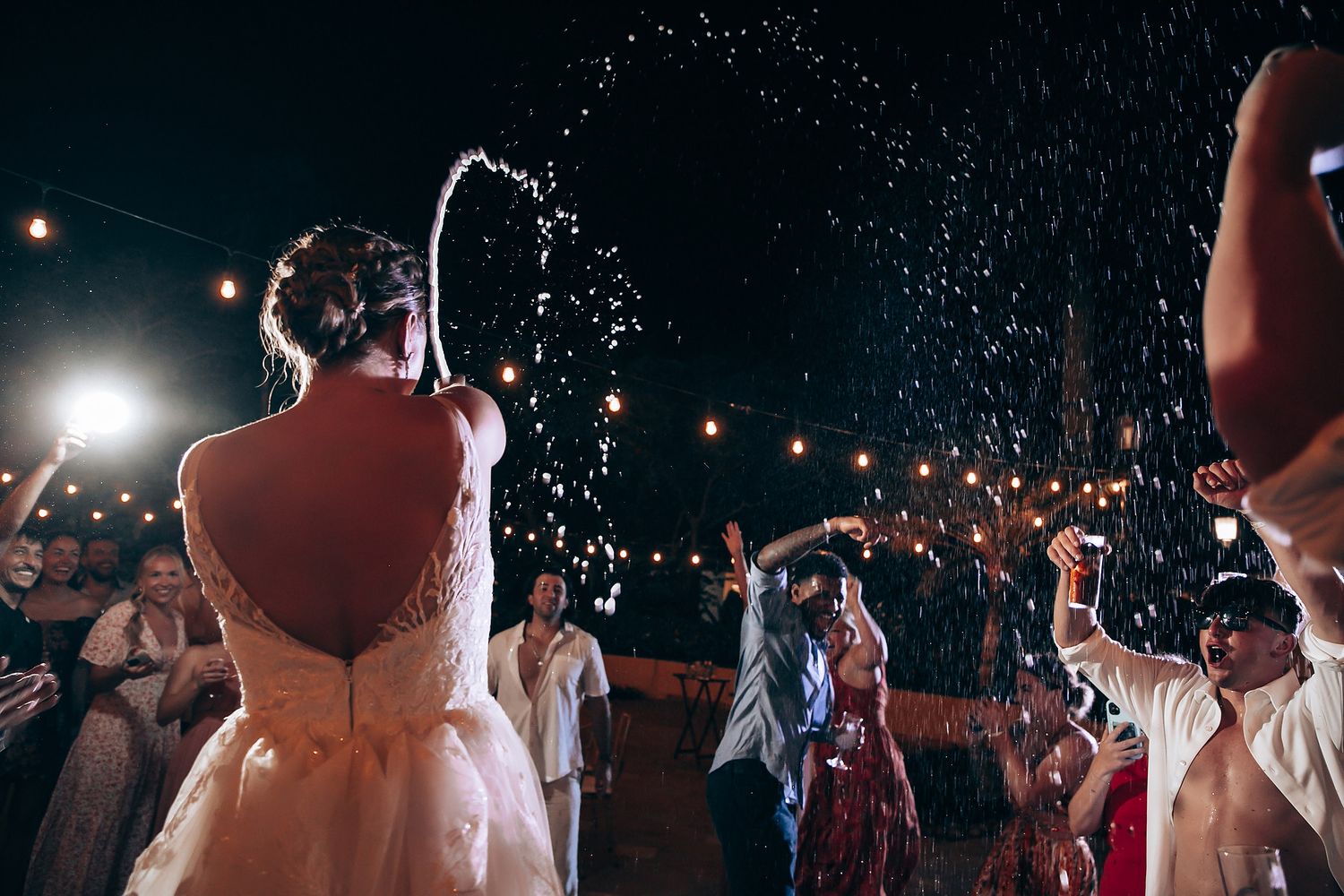 Wedding guests dance and celebrate under twinkling lights as water sprays through the air.
