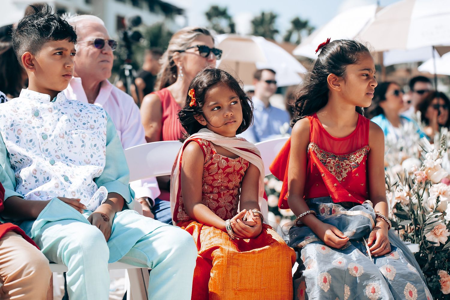 Wedding guests in colorful attire sit outdoors watching a ceremony on a sunny day.