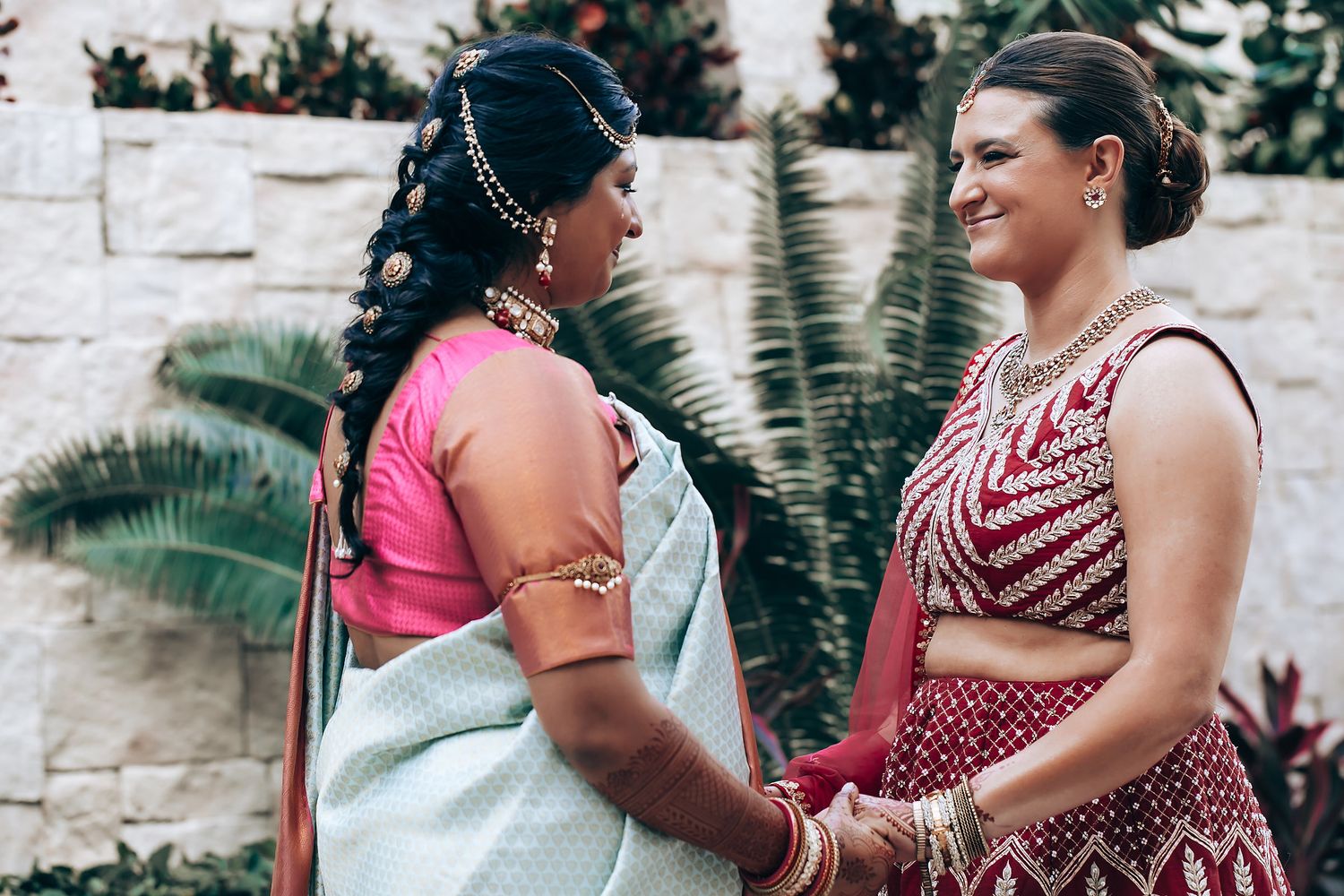 Two people wearing traditional South Asian wedding attire share an intimate moment against a stone wall backdrop.