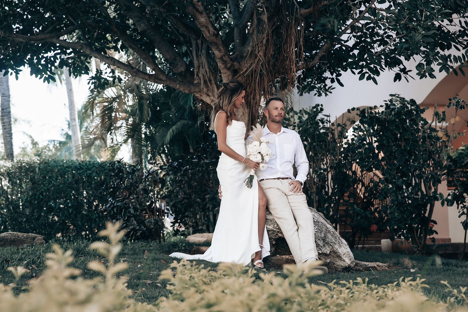 A bride and groom embrace in a lush, green garden setting surrounded by plants and trees.