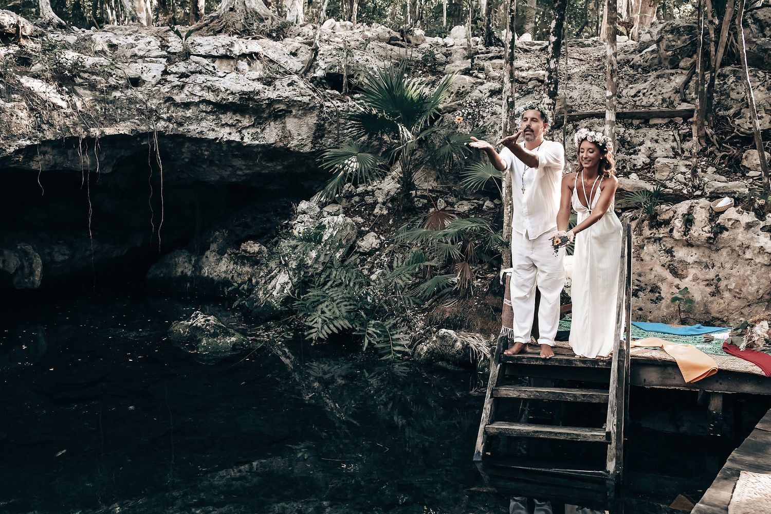 A couple stands on a wooden platform overlooking a lush, forested landscape with a body of water.
