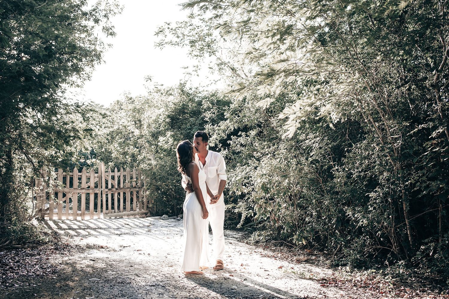 A woman in a white dress jumps on a skateboard in a forested area.