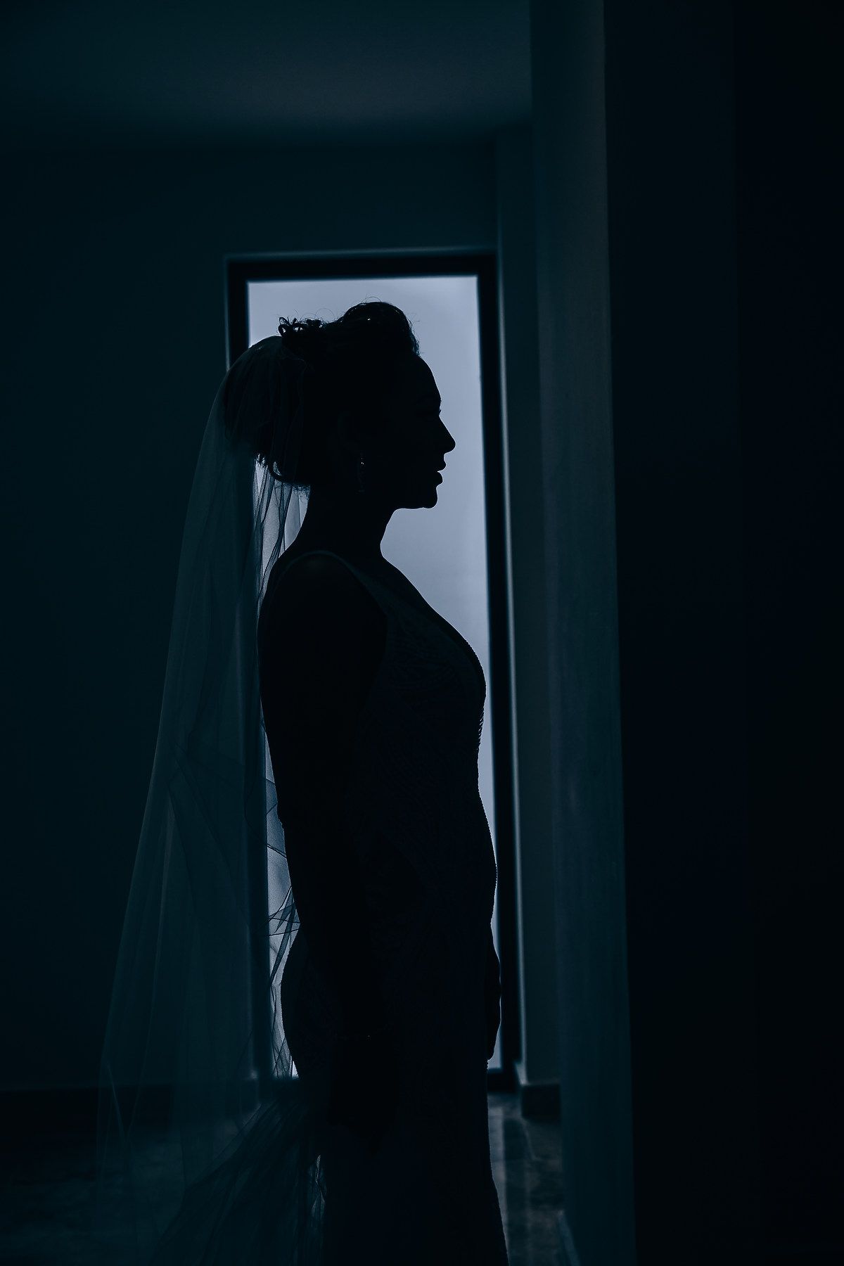 Silhouetted figure stands in dark room against window creating moody atmospheric portrait.