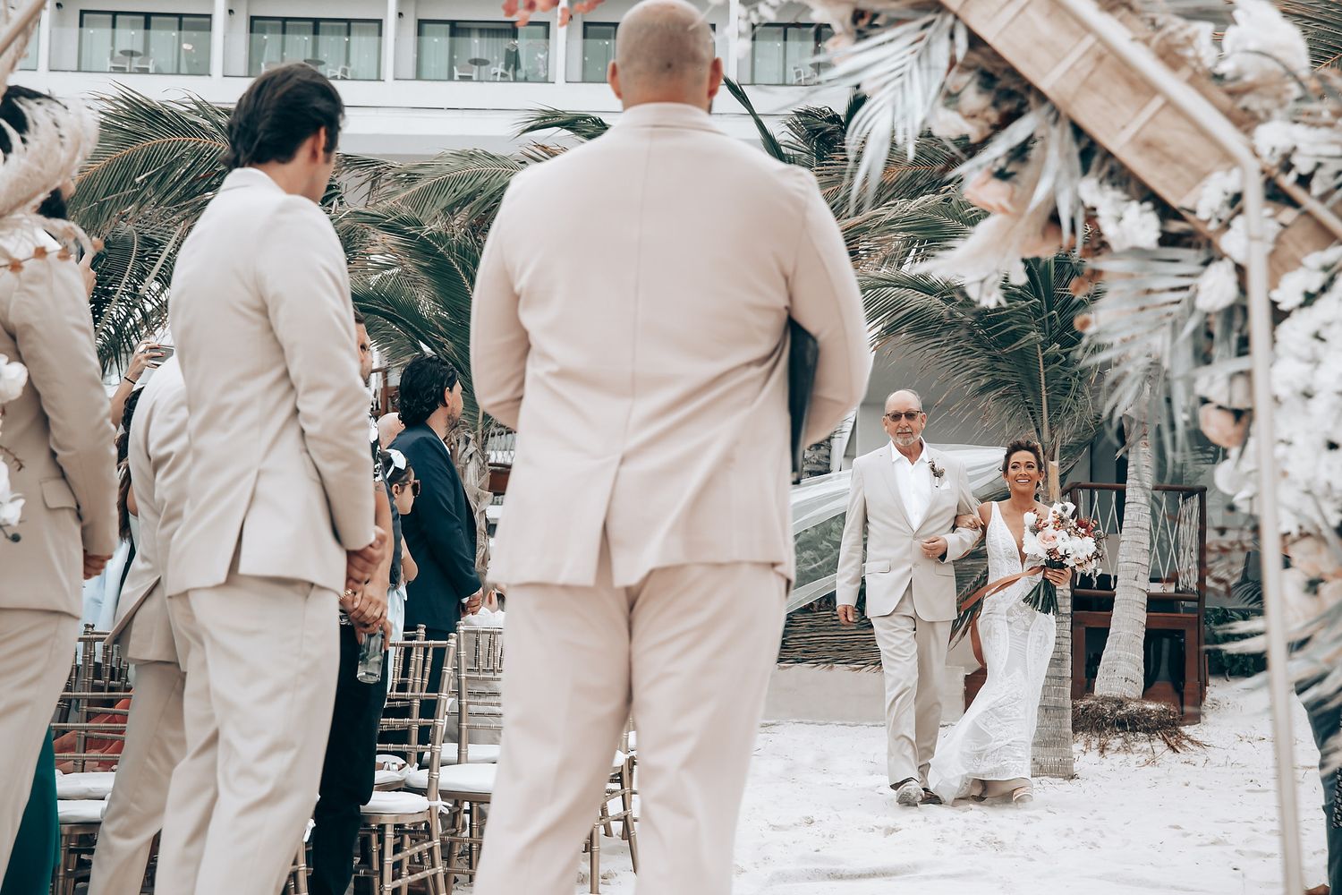 Wedding guests in light beige suits gather for an outdoor beach ceremony.