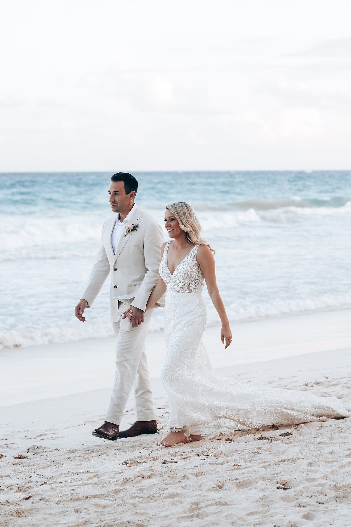 A couple in white wedding attire walks along a pristine beach with turquoise ocean waves.