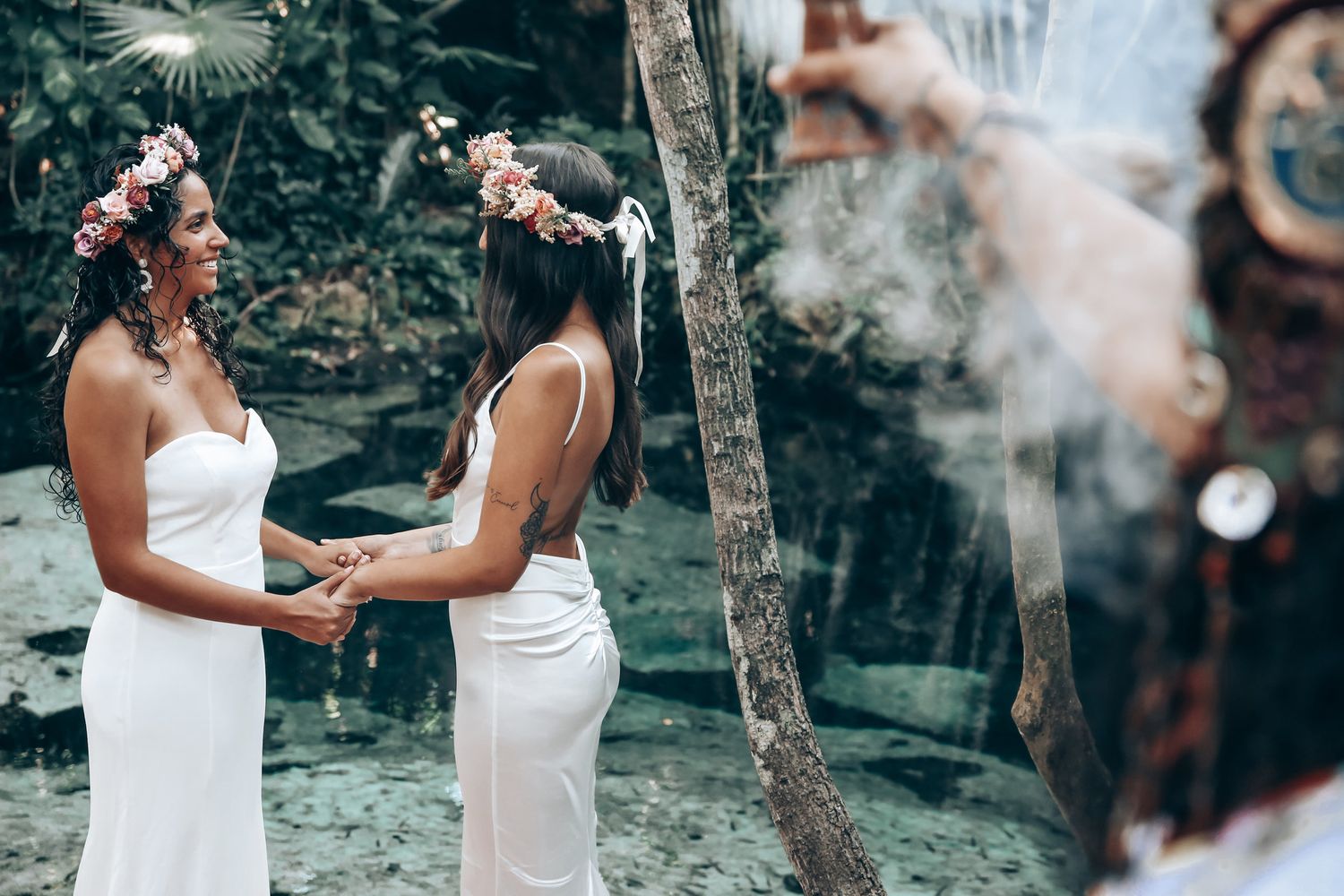 Two people in white dresses hold hands during an outdoor wedding ceremony surrounded by greenery.