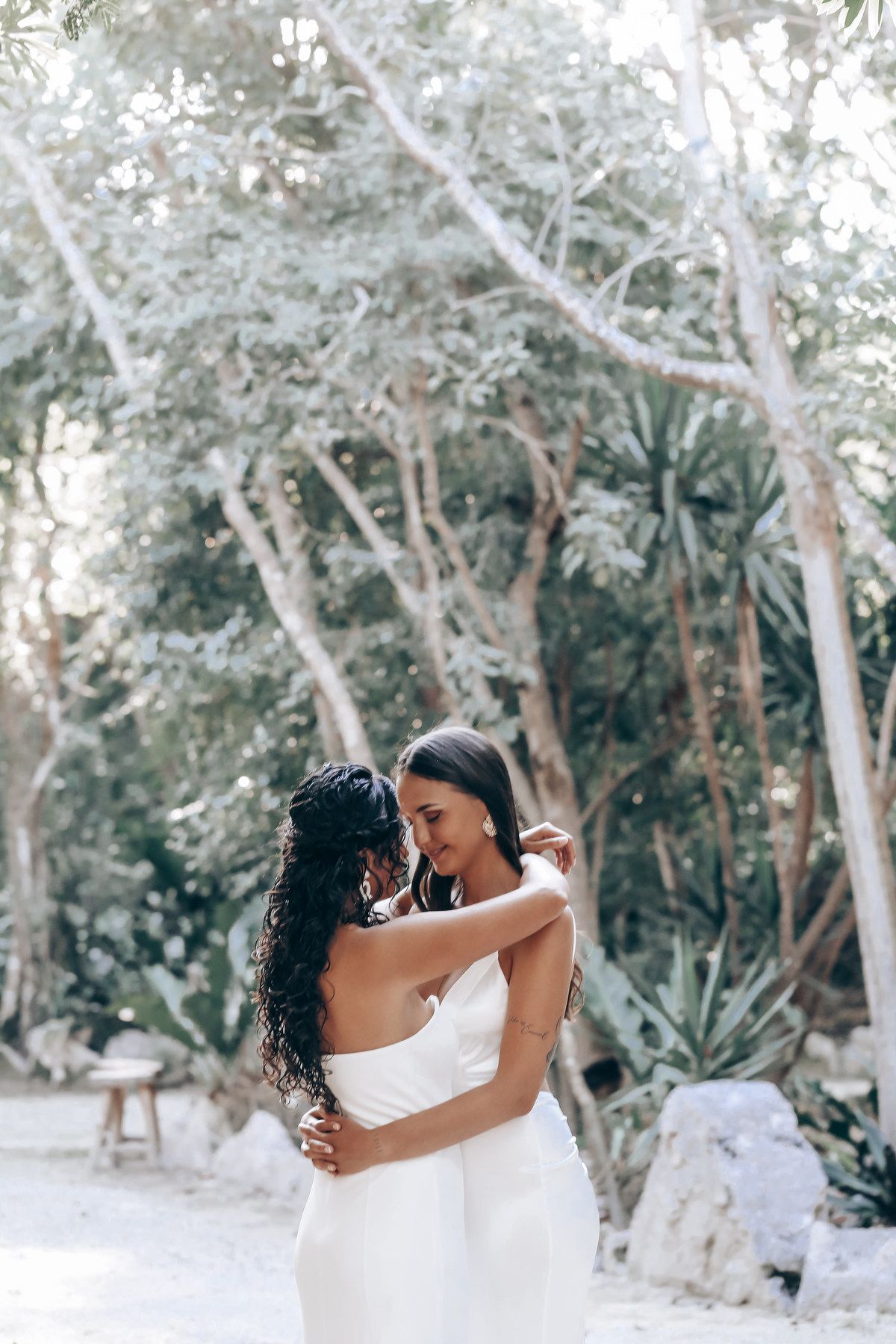 Two people in white outfits share an intimate moment in a sunlit forest setting.
