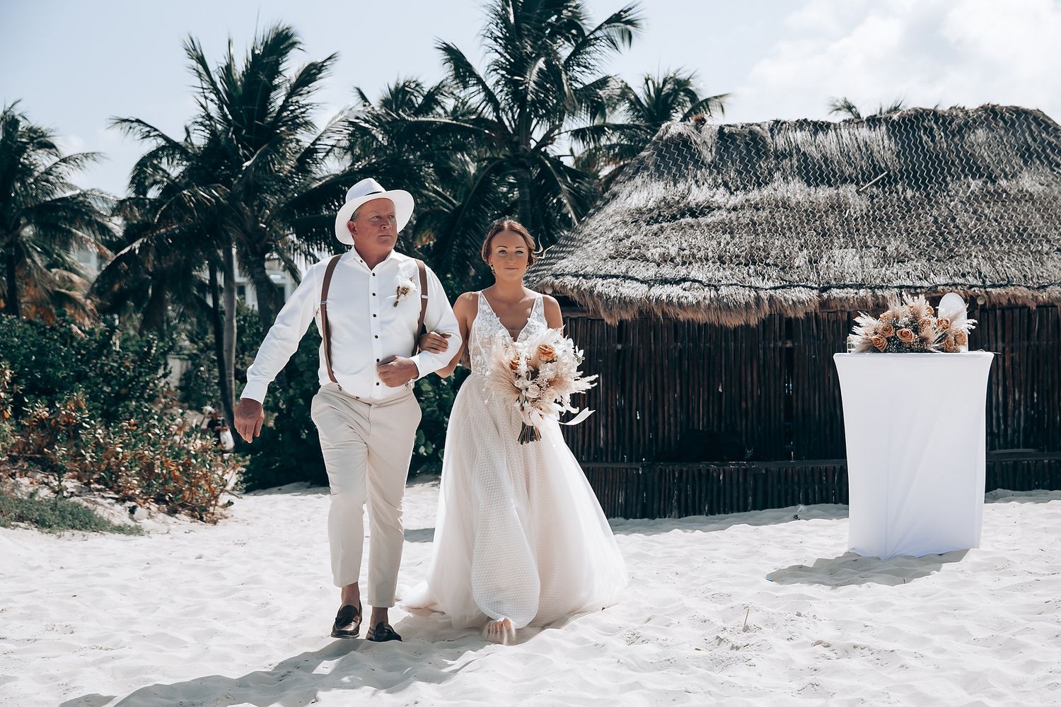 A beachside wedding ceremony with palm trees and thatched roof structures in the background.