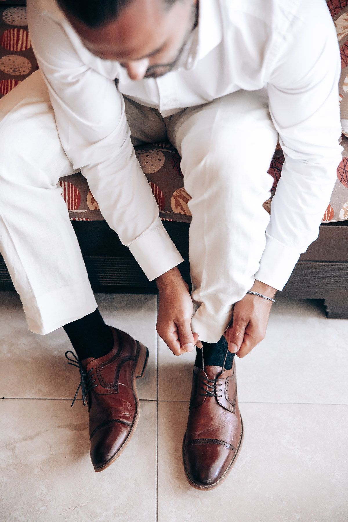 A person in white formal attire tying brown leather dress shoes while sitting on a bench.
