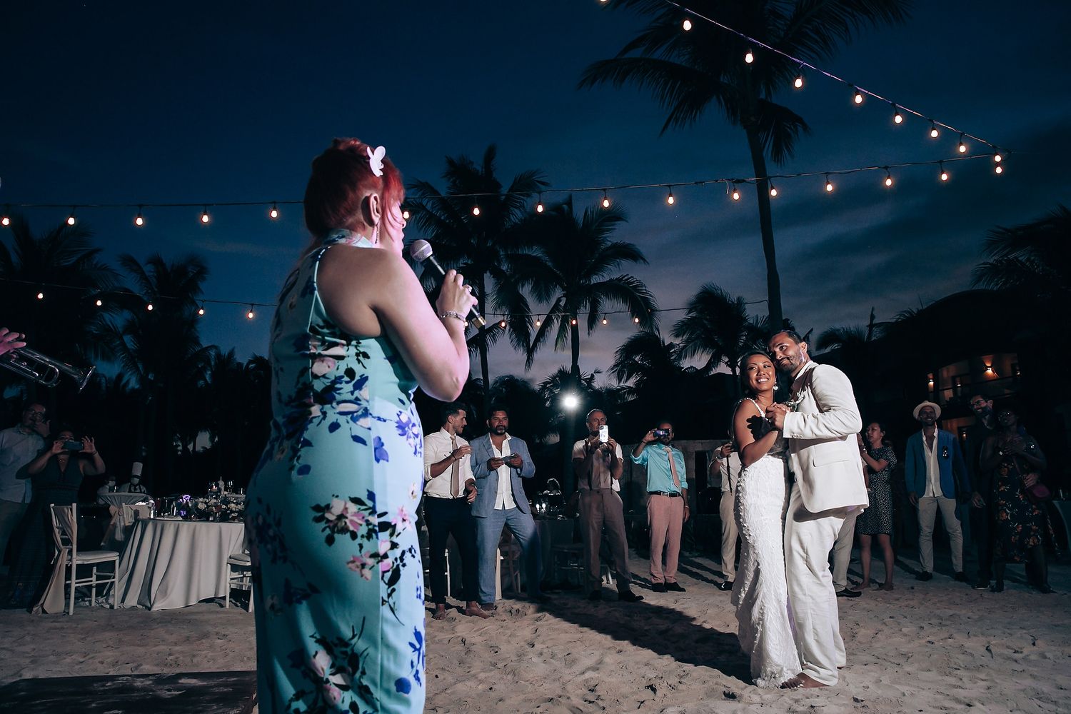 Evening beach wedding reception with string lights and guests mingling under palm trees.