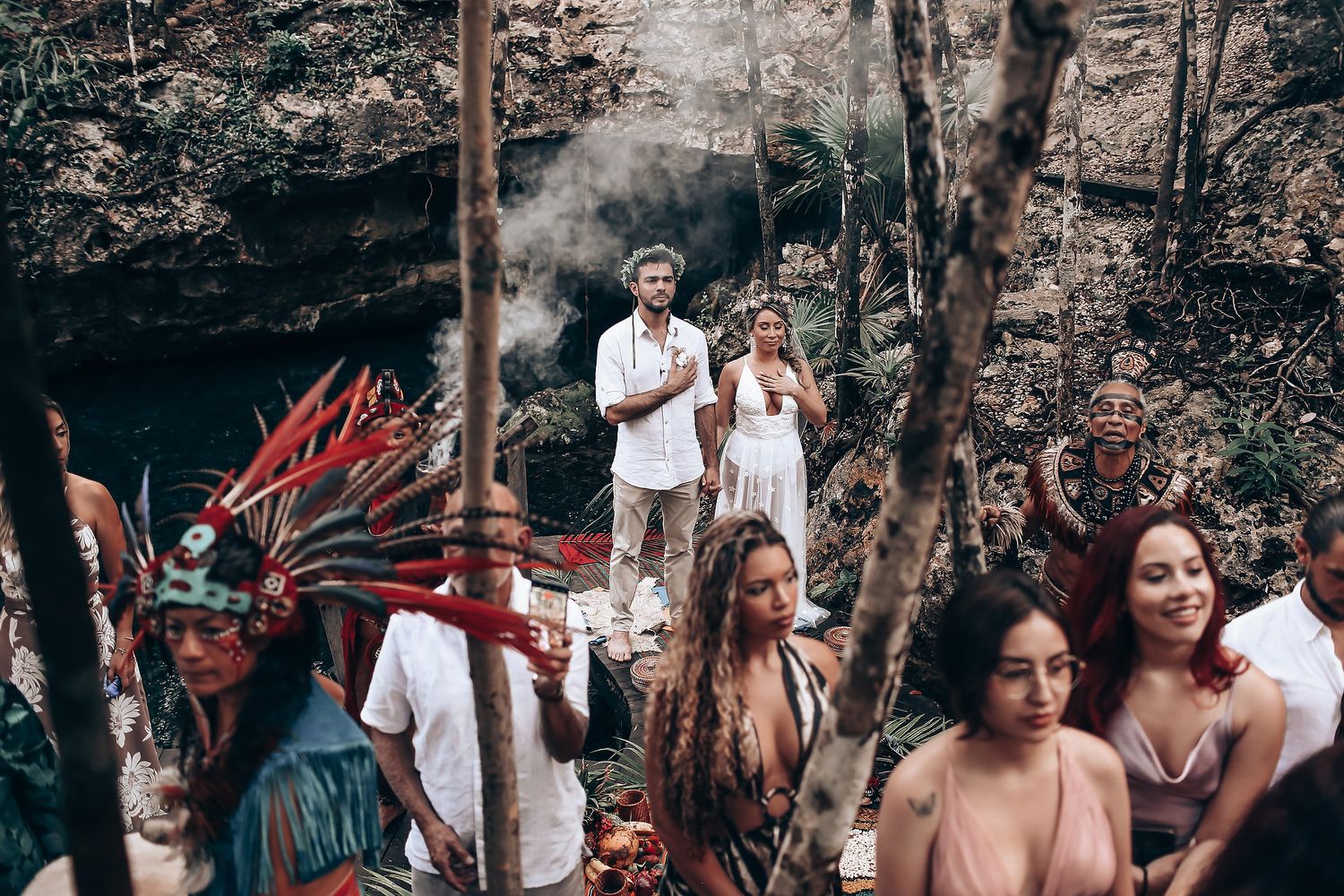 People in white clothing stand among trees during what looks like a forest gathering or celebration.