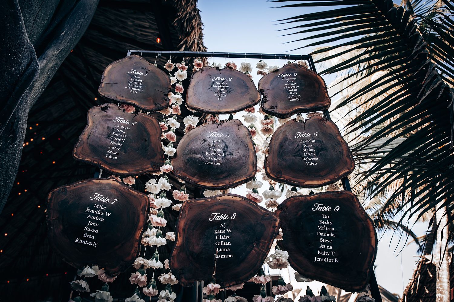 Wedding seating chart display with dark wooden boards decorated with white flowers and palm leaves.