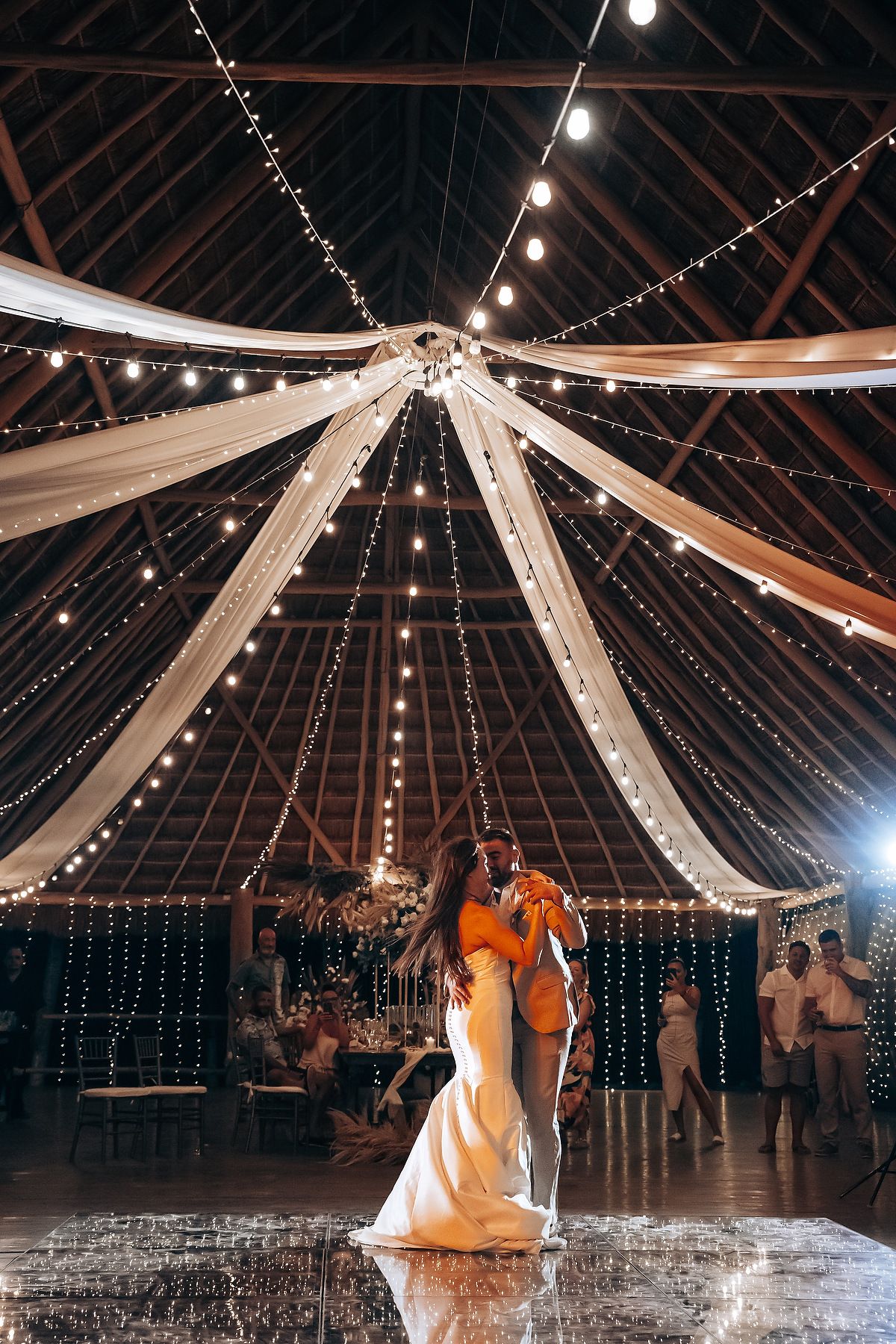 String lights and white fabric drape elegantly from the ceiling of a rustic barn venue creating a romantic atmosphere for dancing.