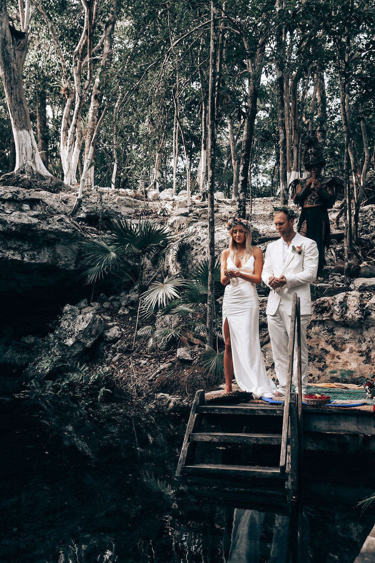 A couple in white wedding attire stands on wooden steps overlooking a cenote in a lush forest setting.