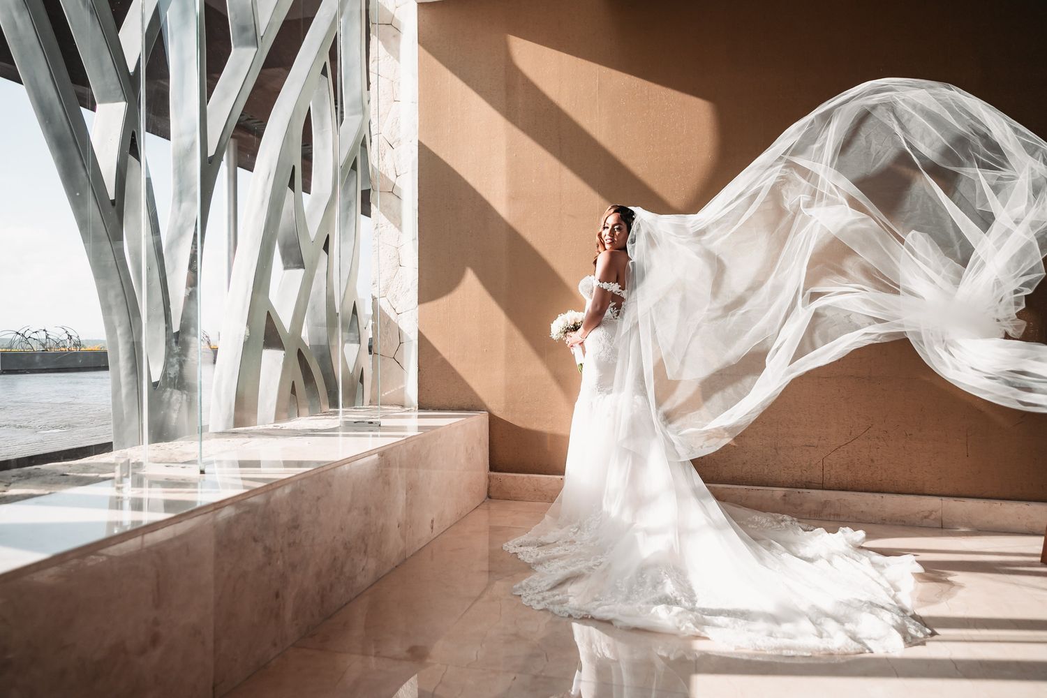 Bride in flowing white gown with dramatic cathedral veil billowing in sunlit architectural space.
