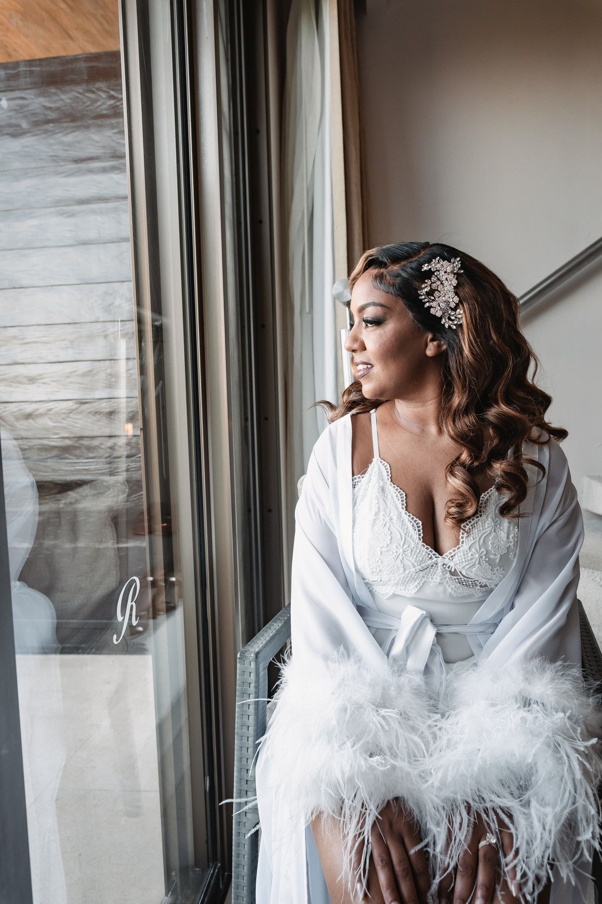 Elegant bride in white feather robe and crystal hair accessory gazing out a window on her wedding day.