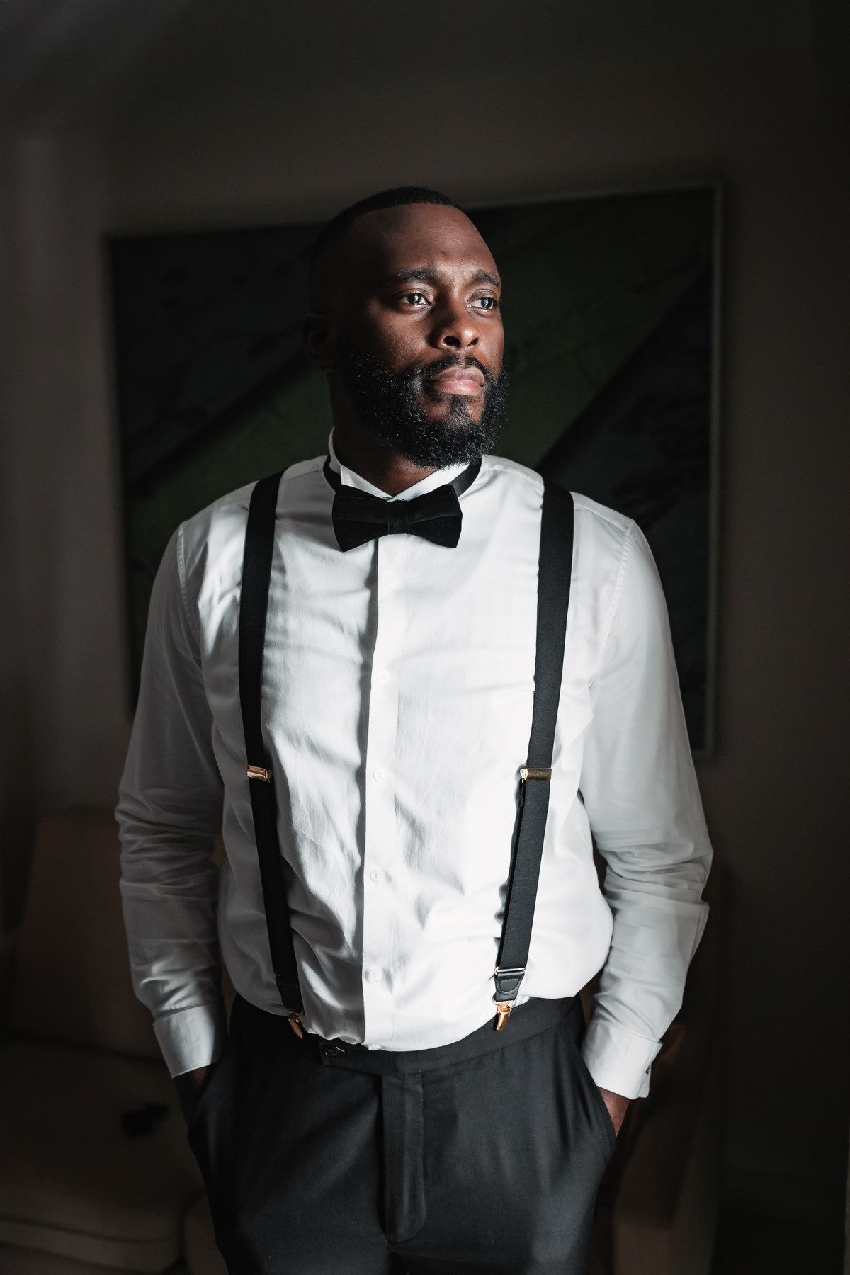Dapper groom in white shirt, black bow tie and suspenders posing indoors.