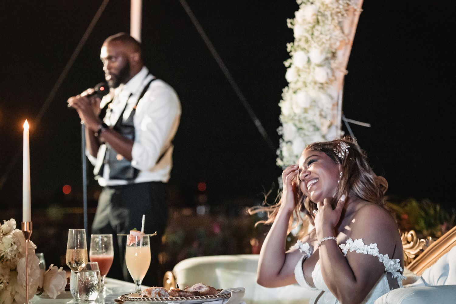 Bride smiling emotionally at sweetheart table while guest performs with violin at outdoor night reception.