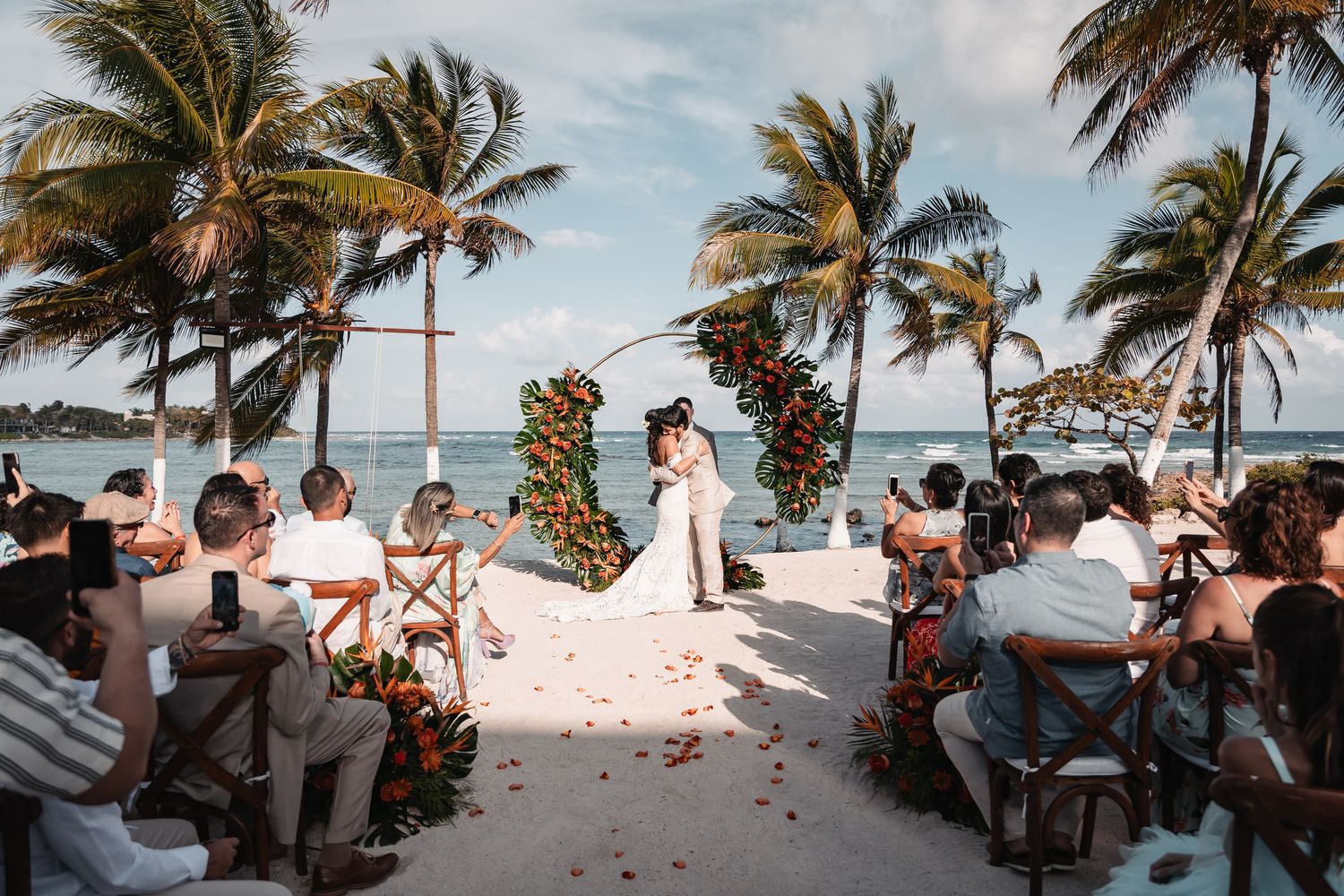 Couple kissing at a romantic beach wedding ceremony with floral arch, palm trees, and ocean backdrop.