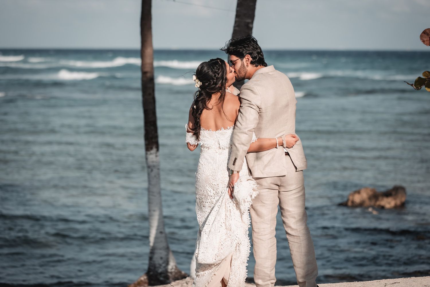 Newlyweds sharing romantic kiss on rocky beach with palm trees nearby.
