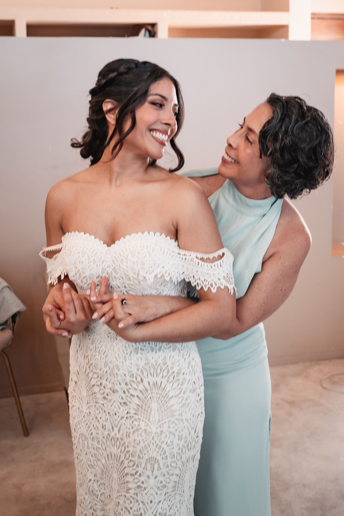 Smiling bride in off-shoulder lace wedding dress being embraced by woman in mint green gown during bridal preparations.