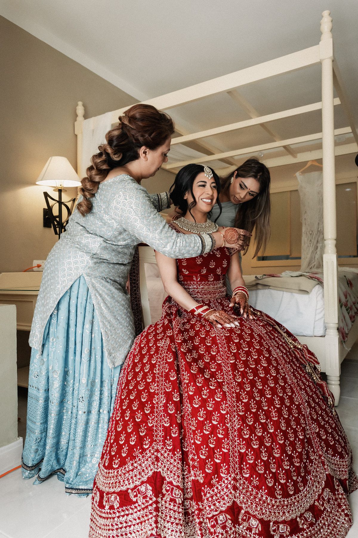 Bride in red lehenga smiles as two women in blue outfits help her prepare for Indian wedding ceremony.