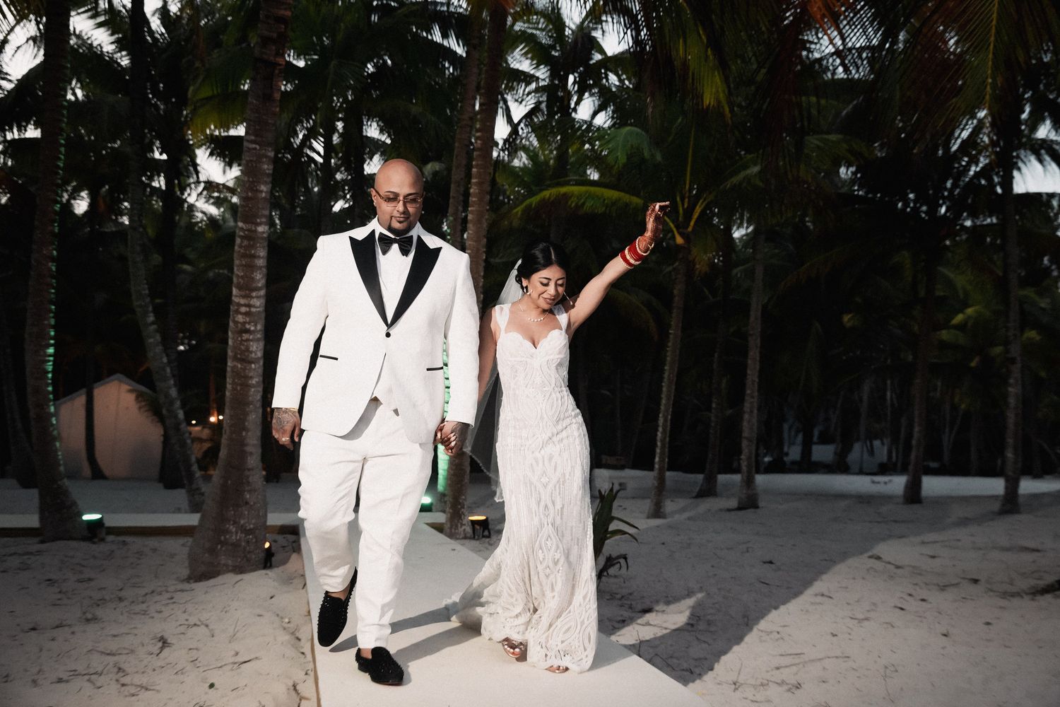Newlywed couple walks hand in hand along sandy beach path lined with tall palm trees after wedding ceremony.