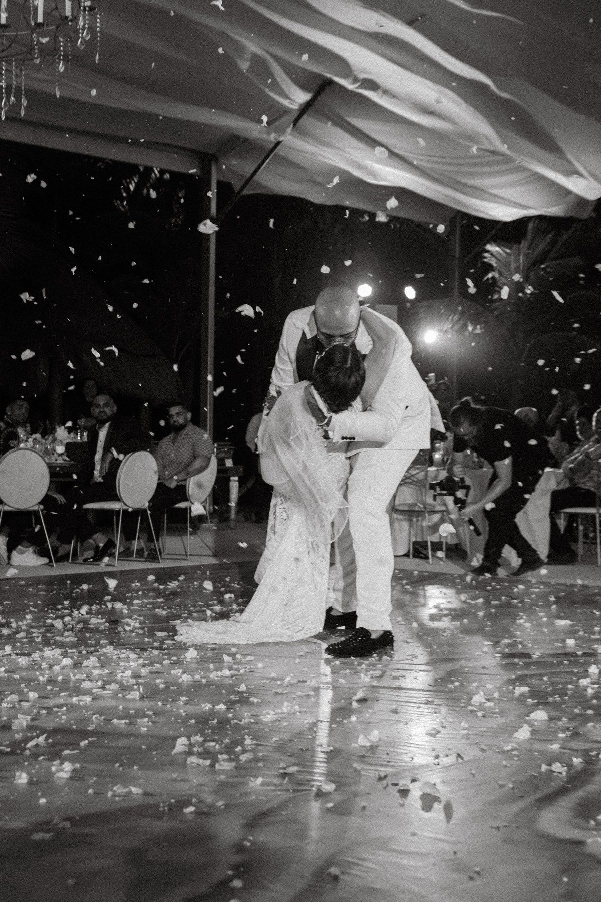 Bride and groom share first dance kiss surrounded by falling flower petals on wedding dance floor in black and white photo.
