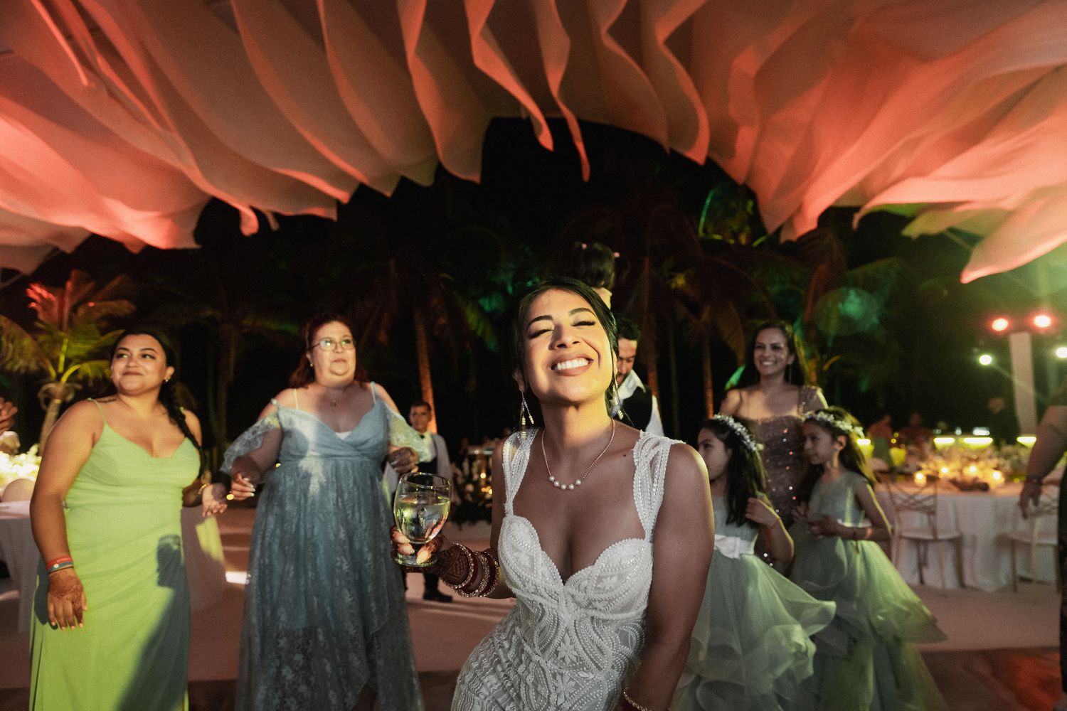 Radiant bride in embellished gown laughs joyfully while dancing with guests at outdoor tropical wedding reception.