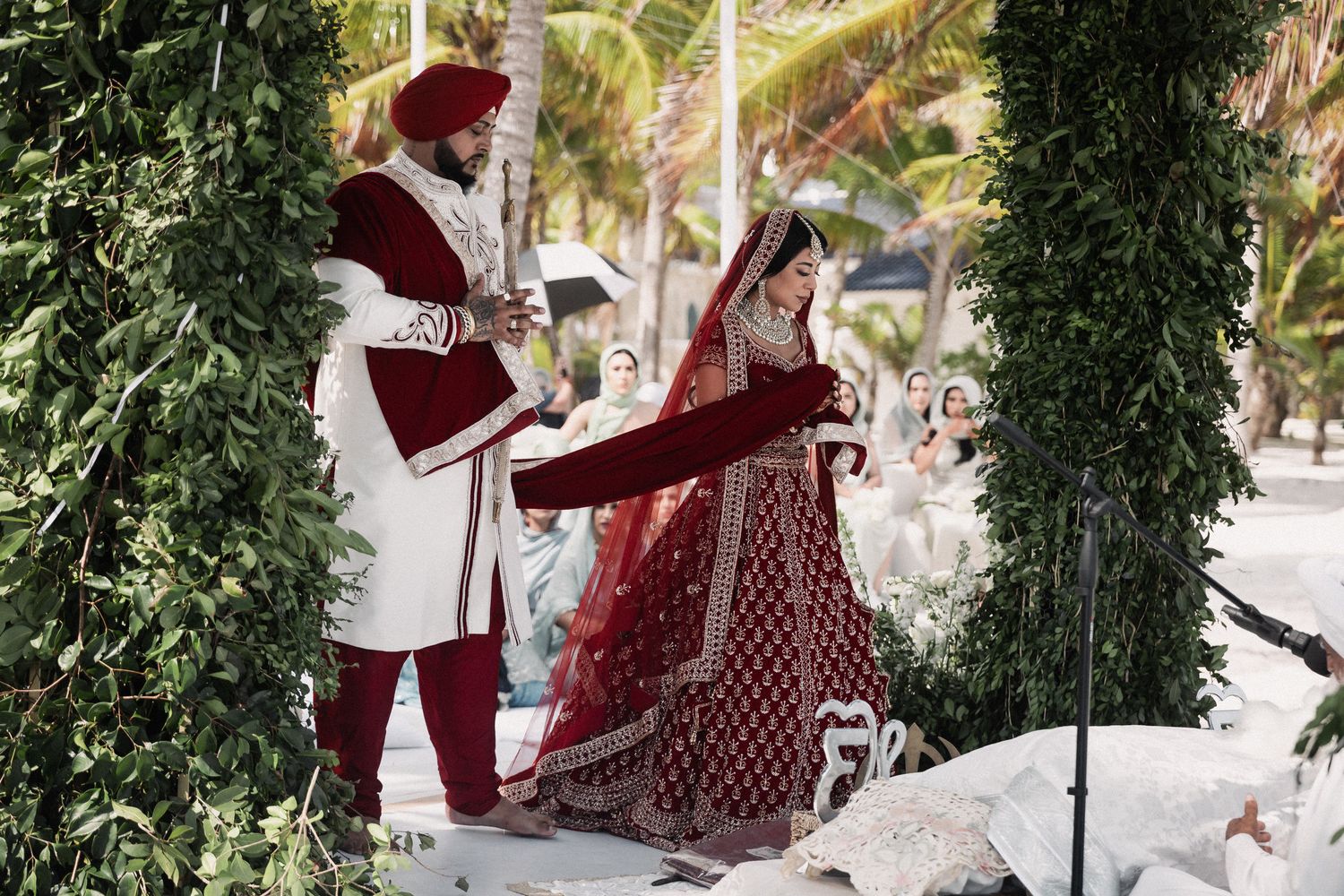 Sikh bride and groom in red and white traditional attire during beach wedding ceremony under floral arch.