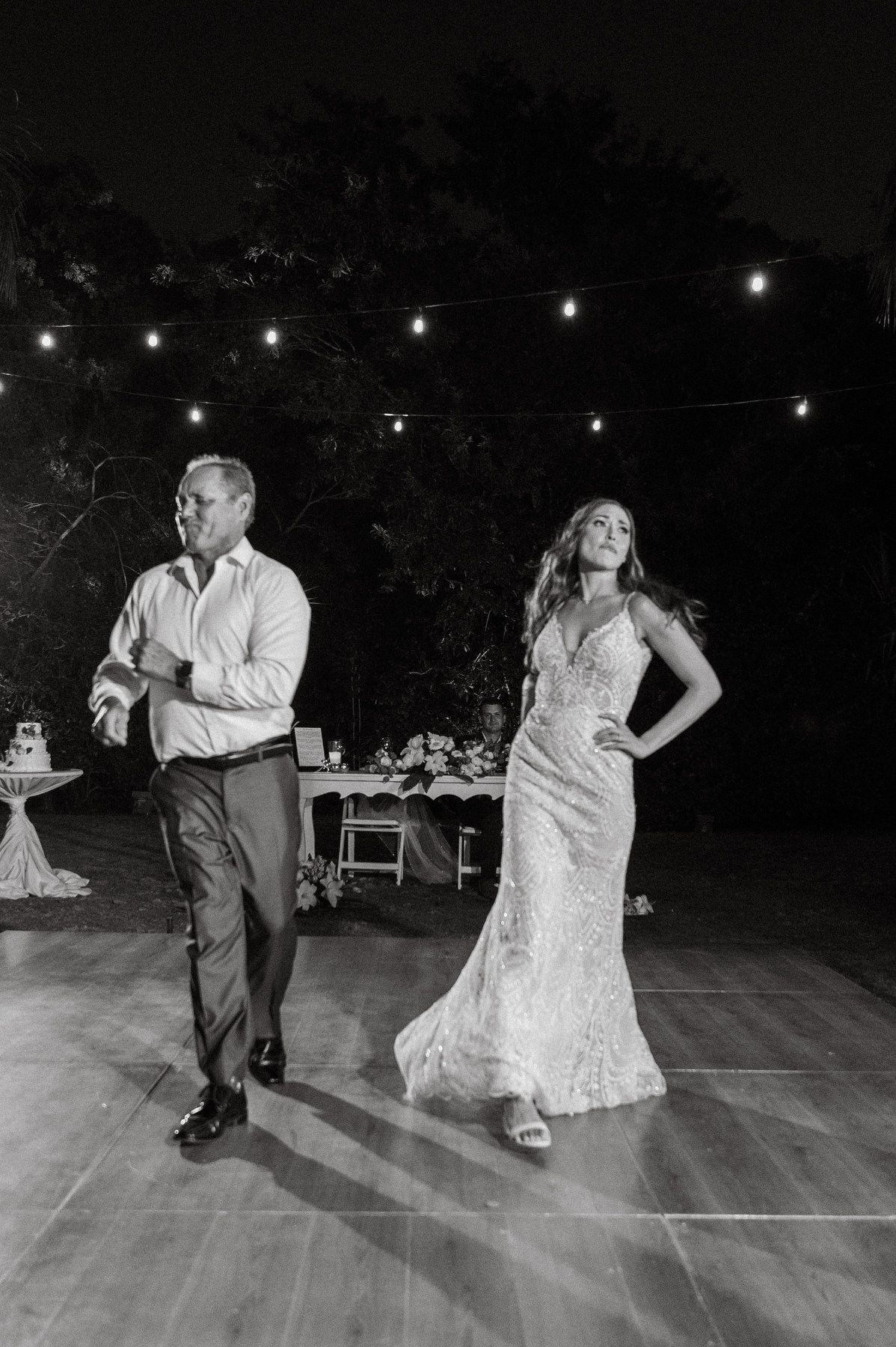 Bride in lace gown and groom dance together on outdoor floor under string lights at a nighttime wedding reception.