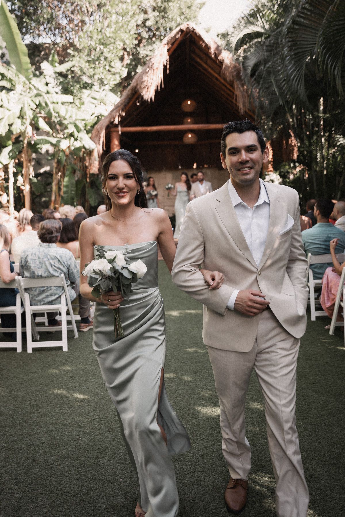 Bridesmaid in sage green gown walks down aisle with groomsman after ceremony.