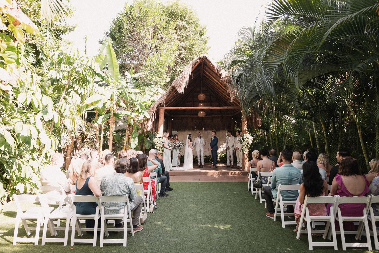 Outdoor wedding ceremony with guests seated in white chairs before rustic thatched-roof altar in tropical garden.