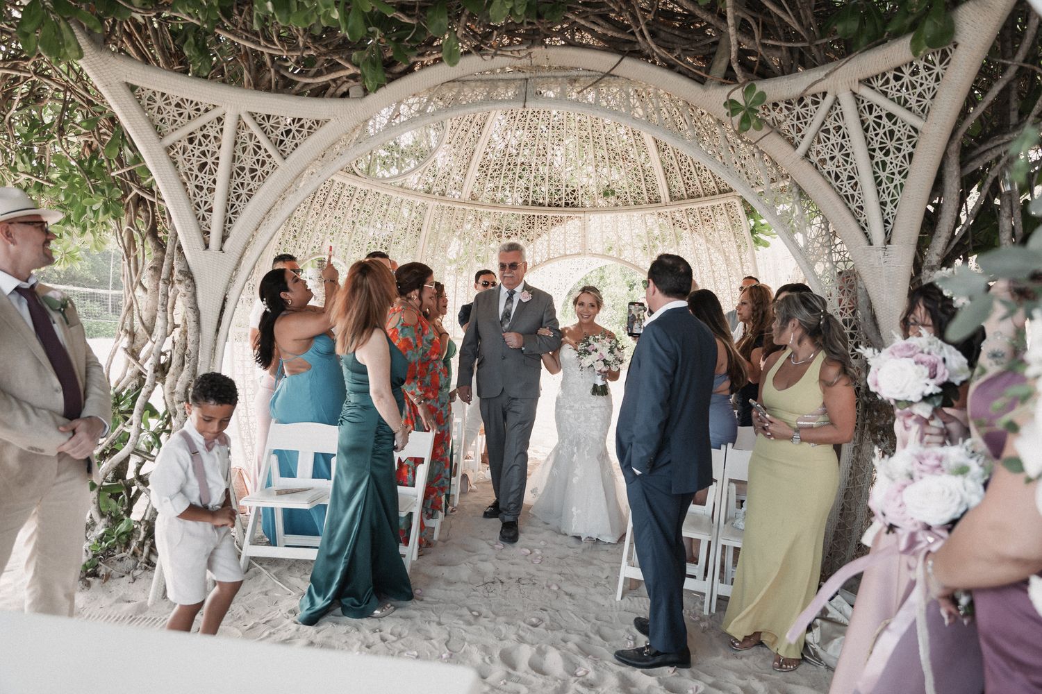 Outdoor beach wedding ceremony under ornate white gazebo with guests seated on white chairs.