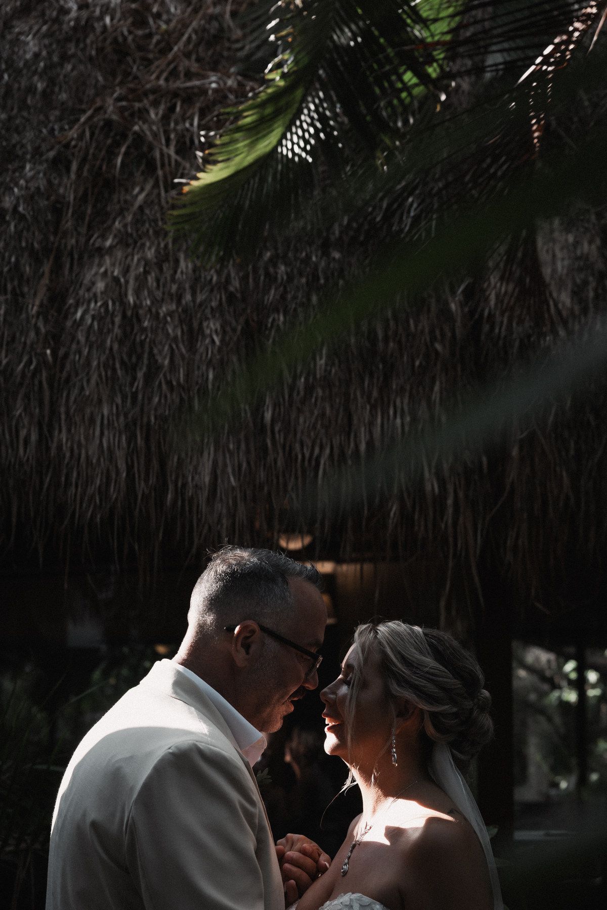 Bride and groom share intimate moment under thatched palapa roof in dramatic tropical lighting.