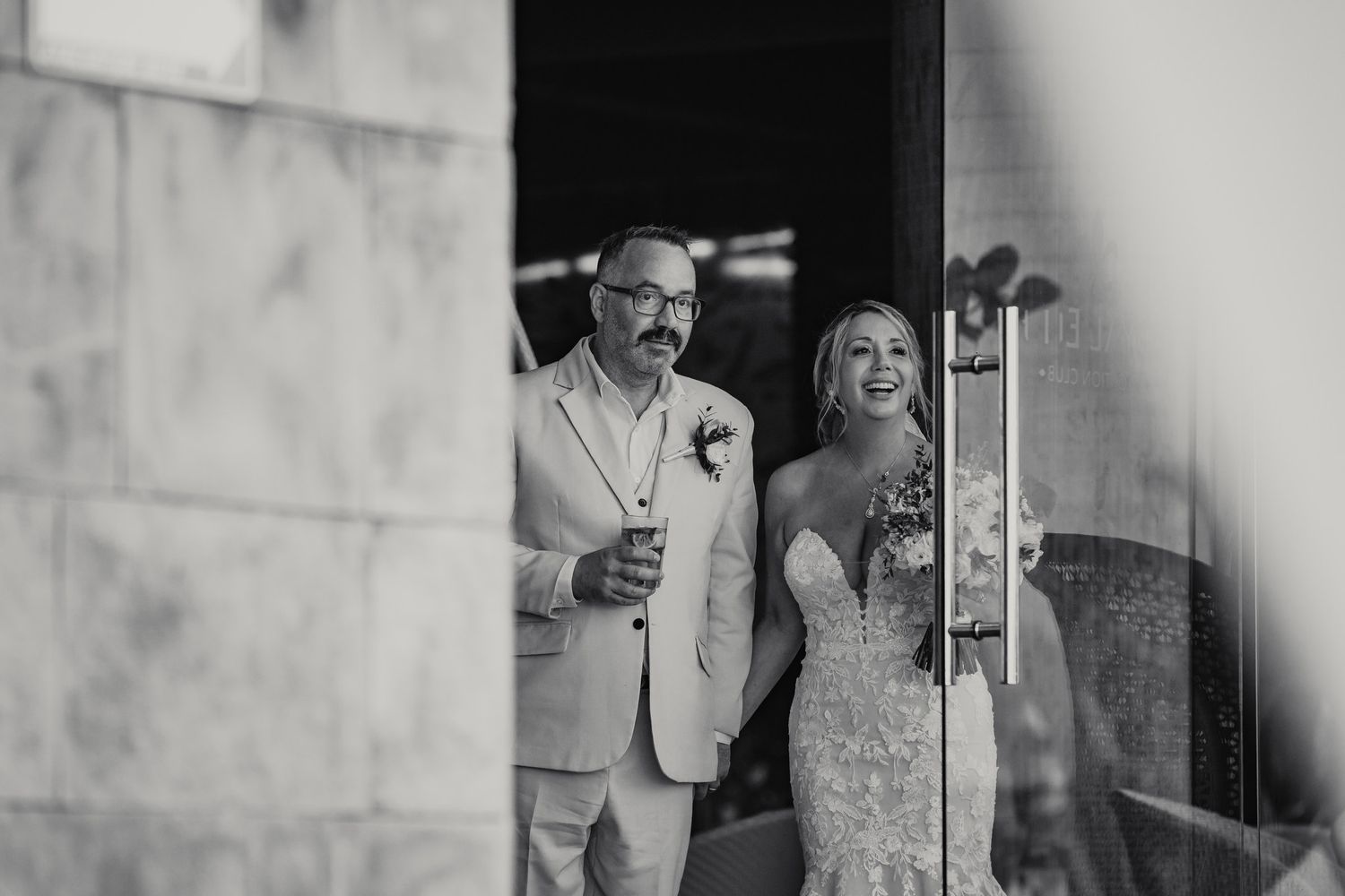 Black and white photo of smiling bride and groom emerging through doorway holding drinks.