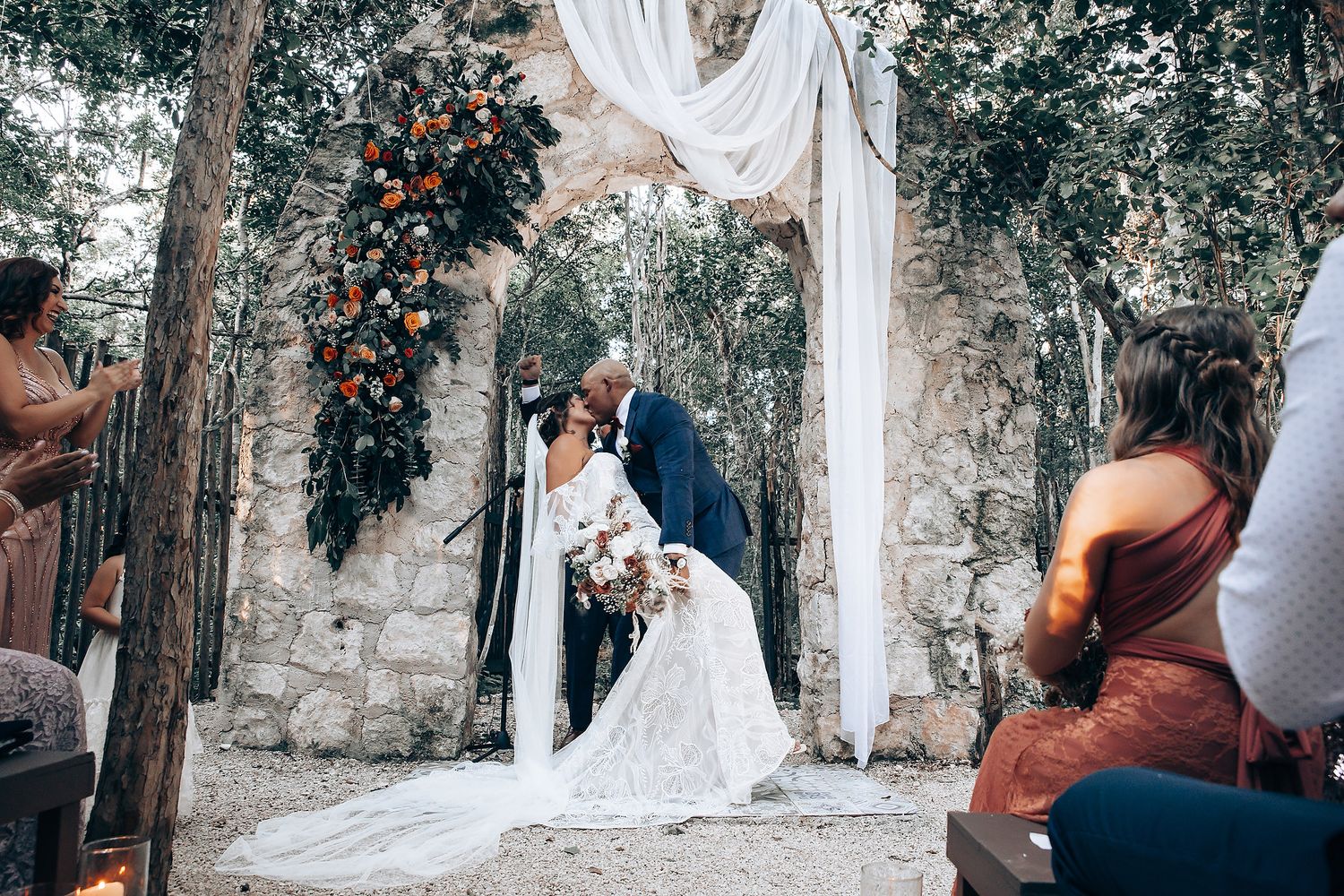 A wedding ceremony under a draped white arch with stone walls and floral decorations.