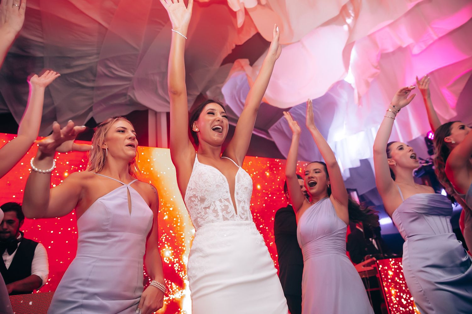 Wedding guests dancing with raised hands under pink and red lighting effects.