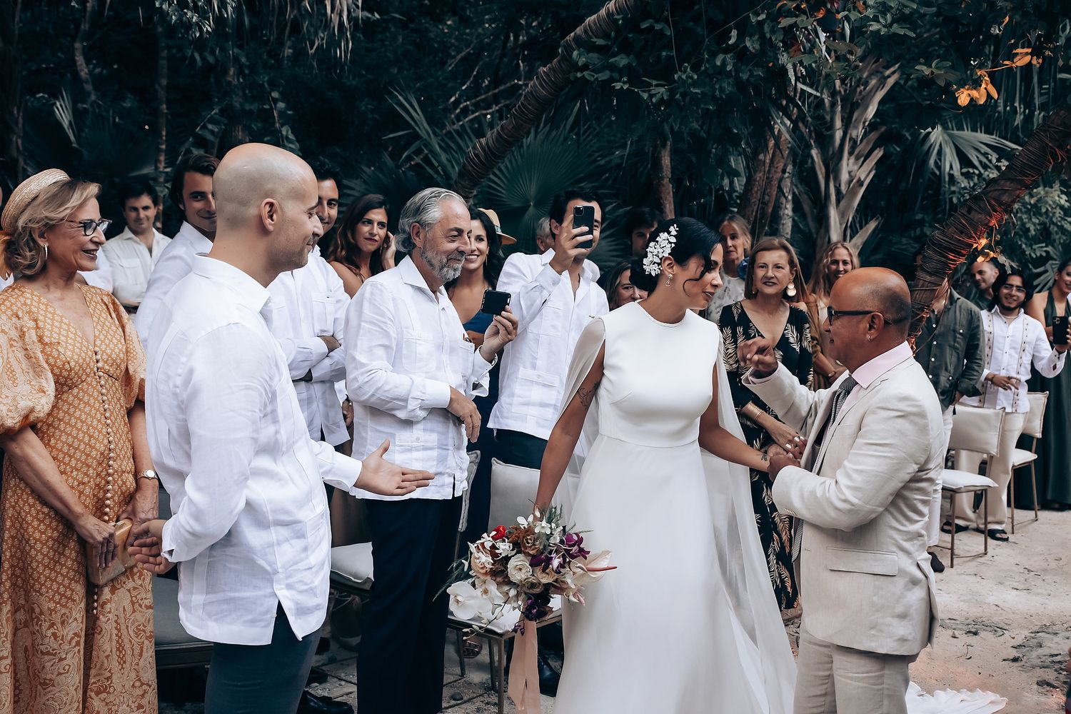 Wedding guests in white and beige attire mingle at an outdoor tropical celebration.
