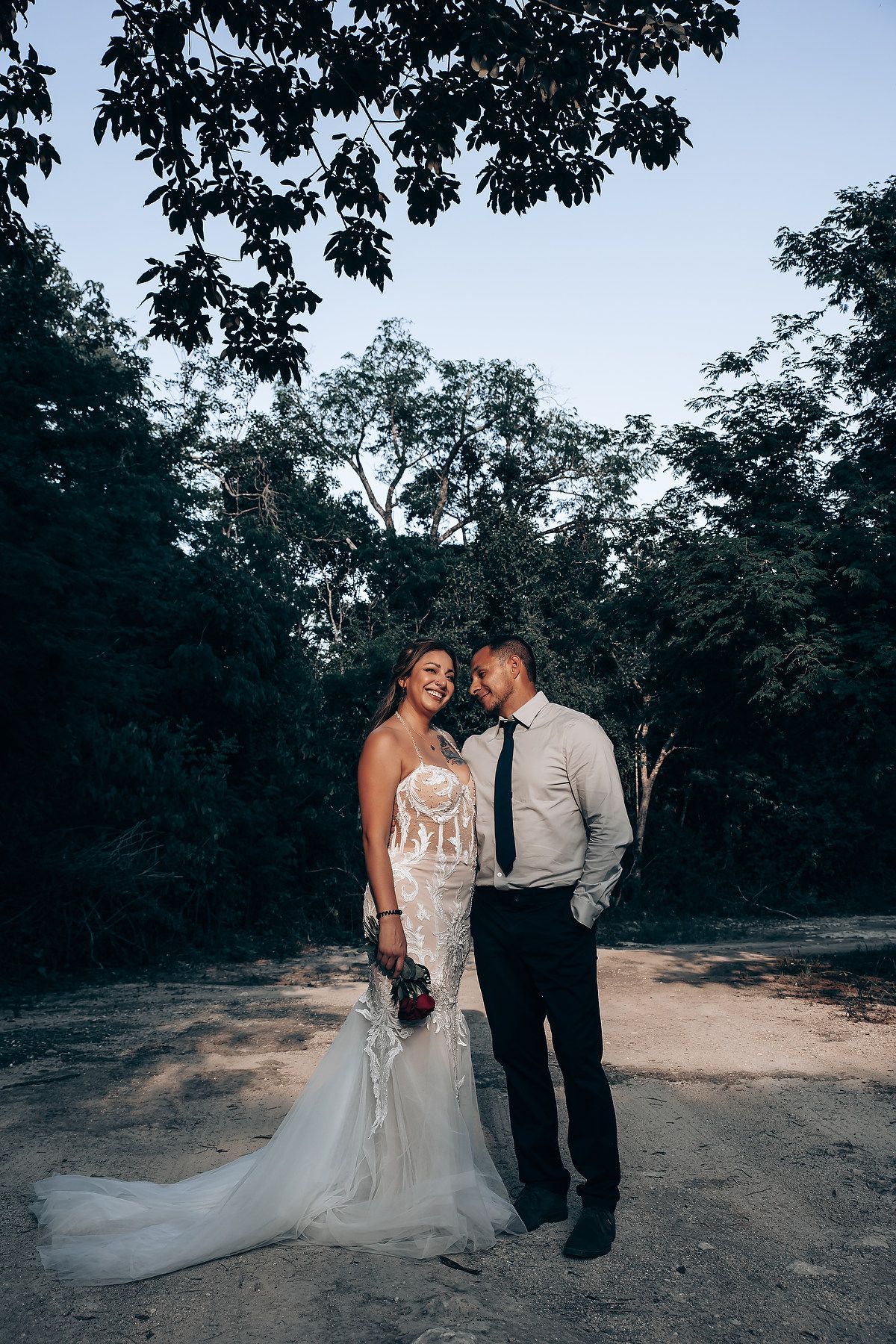 A couple shares an intimate moment in their wedding attire under trees at sunset.