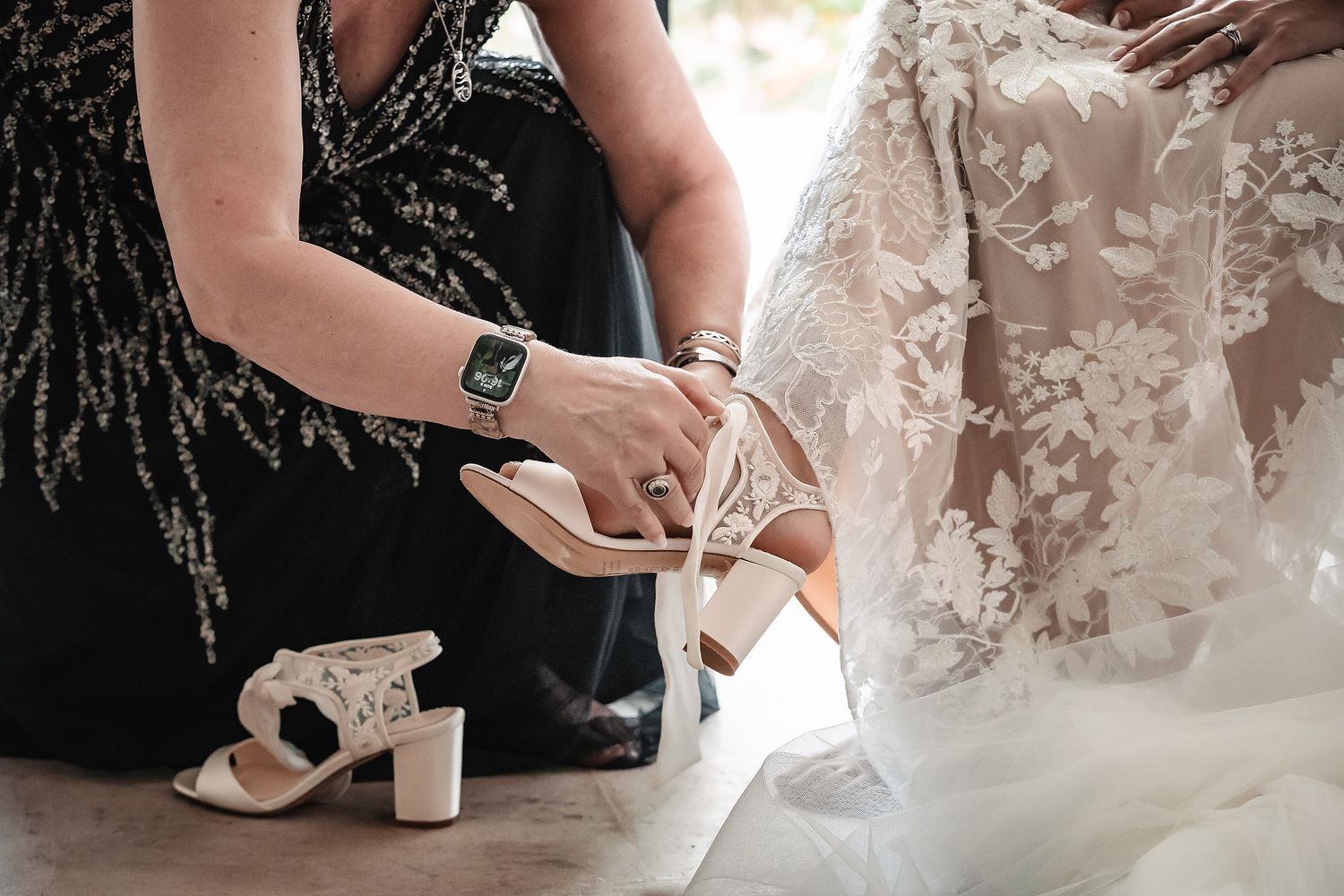 A woman helps a bride put on elegant strappy wedding heels while another white block-heel shoe sits nearby.