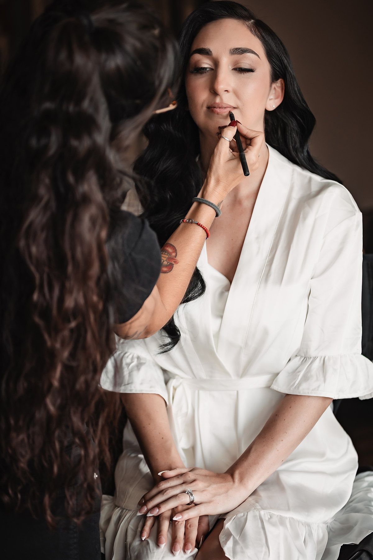 Makeup artist applying lipstick to bride in white satin robe before wedding.