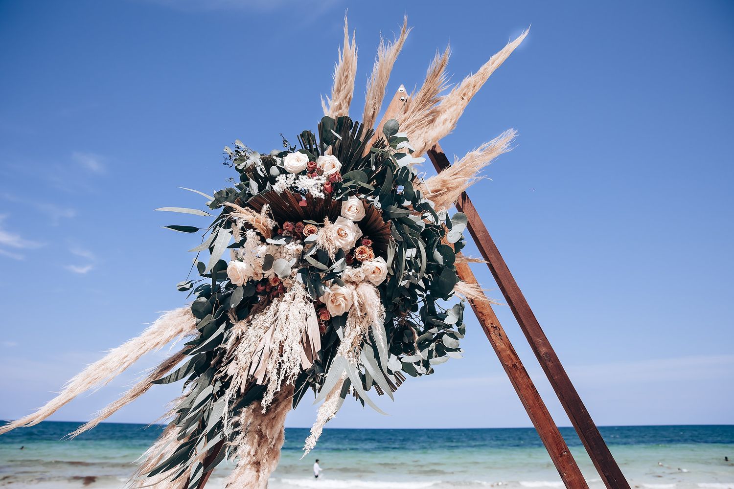 Tropical beach wedding arch decorated with pampas grass and dried palm fronds against turquoise ocean waters.