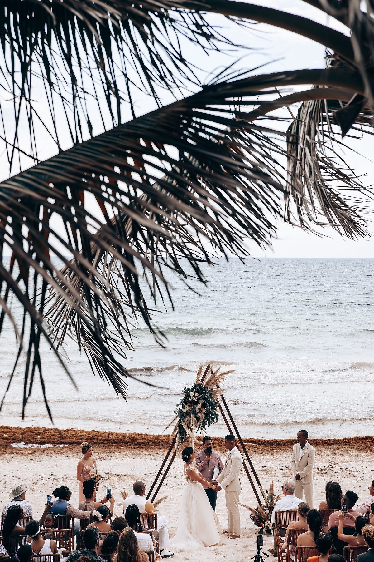 Beachside wedding ceremony under palm trees with guests seated on white chairs facing the ocean.